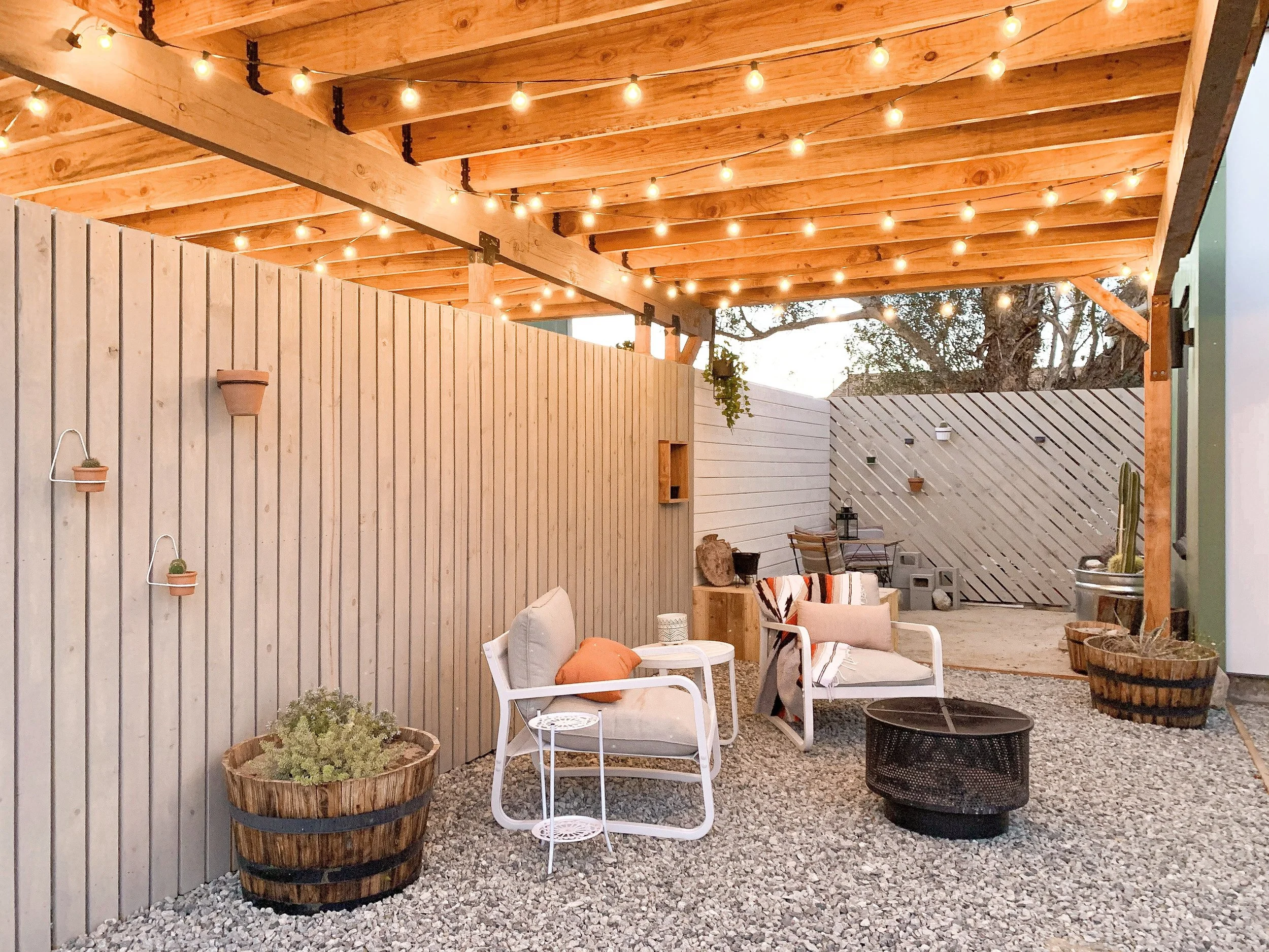 Cozy backyard patio area with string lights, wooden fencing, cushioned chairs, small tables, rustic fire pit, and large planters with plants.