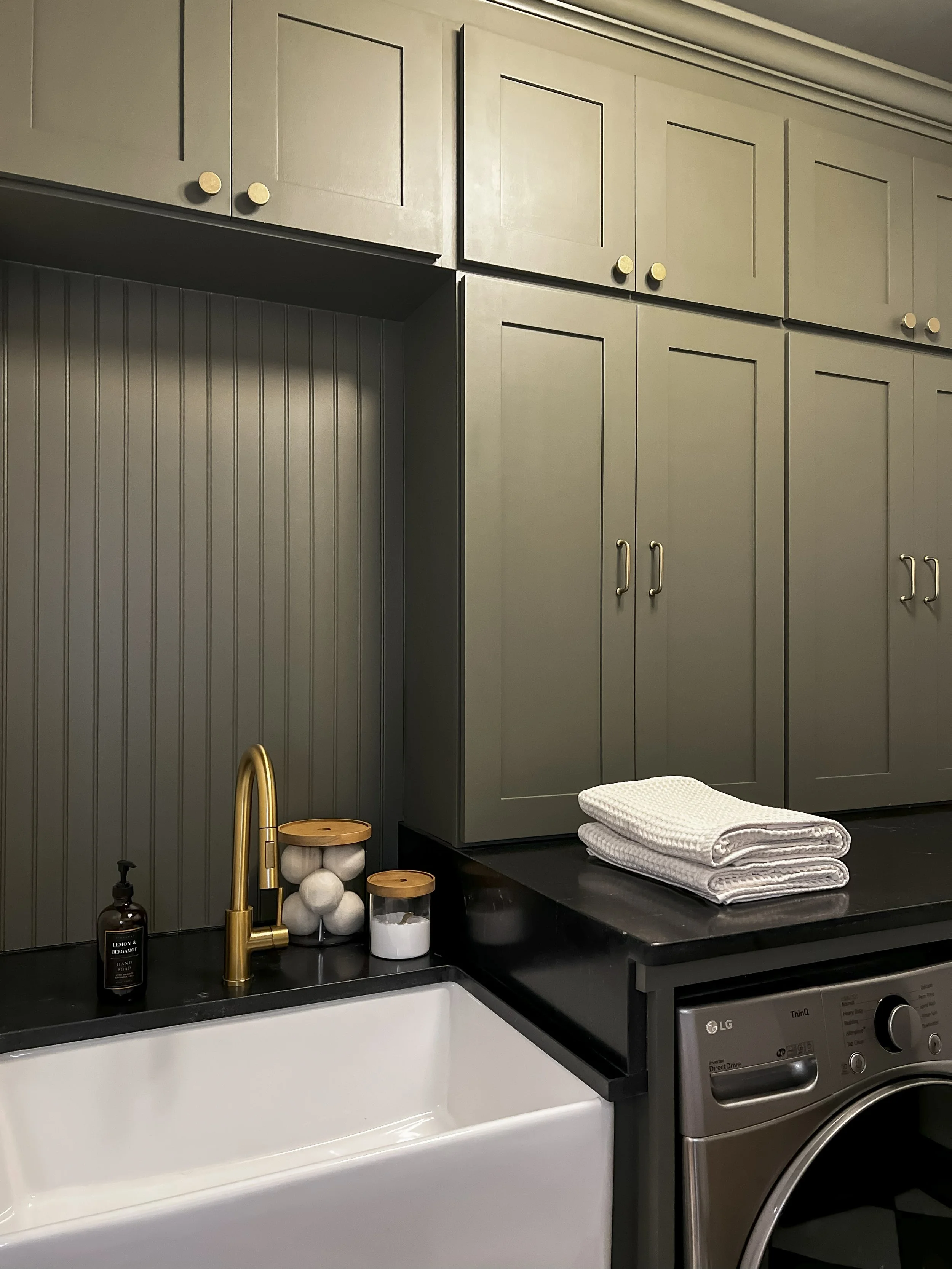 Interior design of an upscale laundry room for a Philadelphia area home. Embodying a modern yet timeless style, this space features dark olive custom cabinets, black granite counter above built-in laundry machines, and checker tile flooring.
