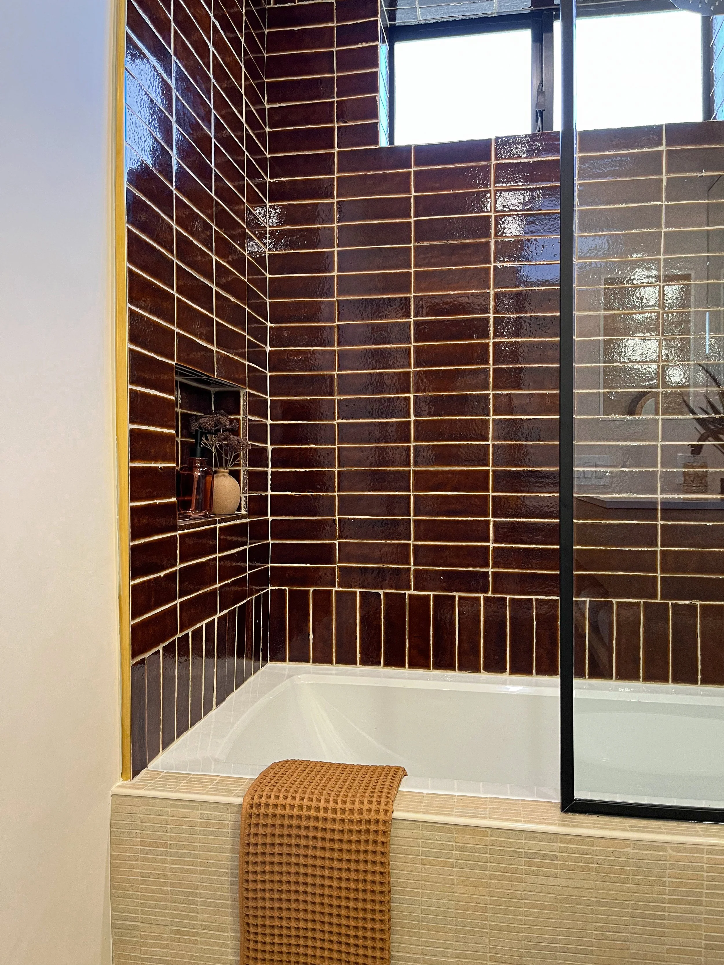 A modern bathroom shower with dark brown glossy tiles, a small built-in shelf with decorative bottles, a clear glass divider, and a brown textured towel hanging over the edge of the bathtub.