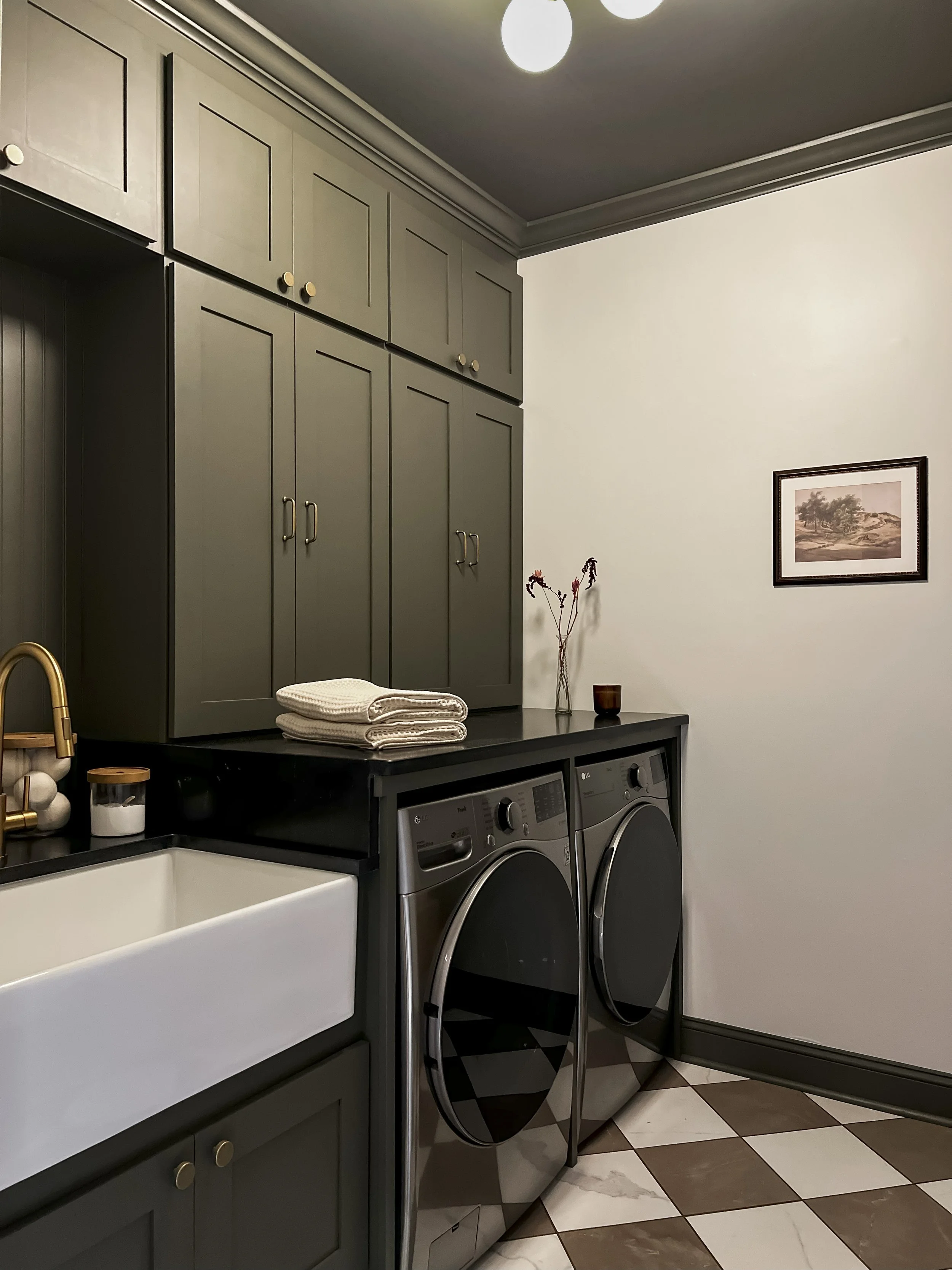 Interior design of an upscale laundry room for a Philadelphia area home. Embodying a modern yet timeless style, this space features dark olive custom cabinets, black granite counter above built-in laundry machines, and checker tile flooring.