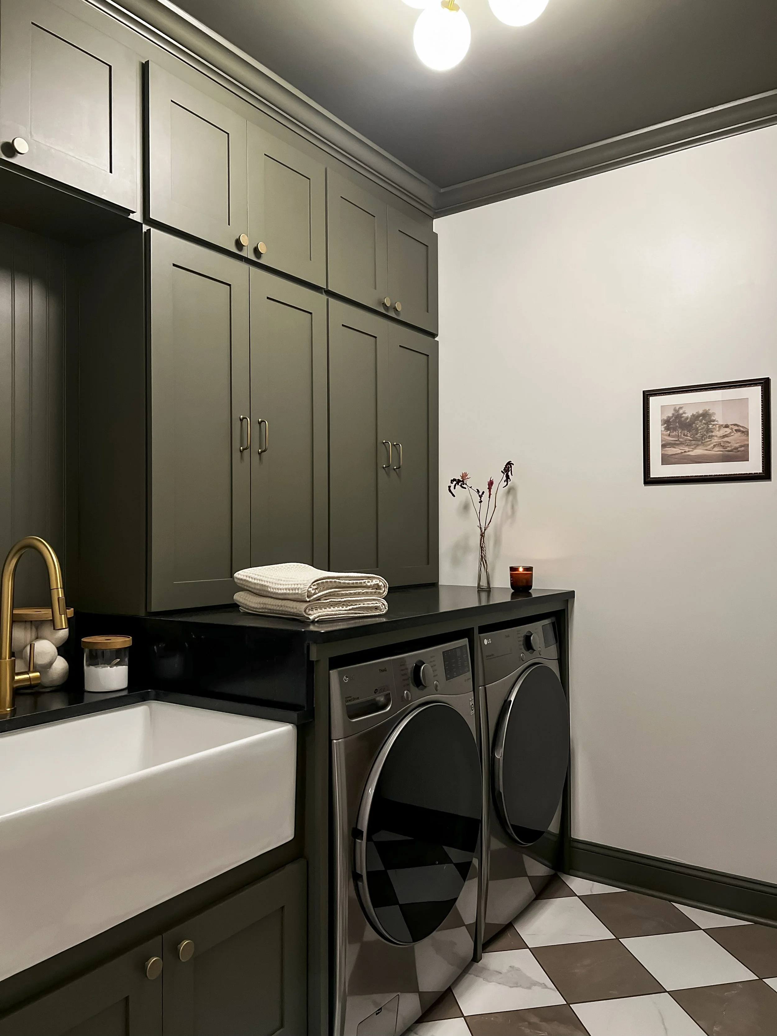Interior design of an upscale laundry room for a Philadelphia area home. Embodying a modern yet timeless style, this space features dark olive custom cabinets, black granite counter above built-in laundry machines, and checker tile flooring.