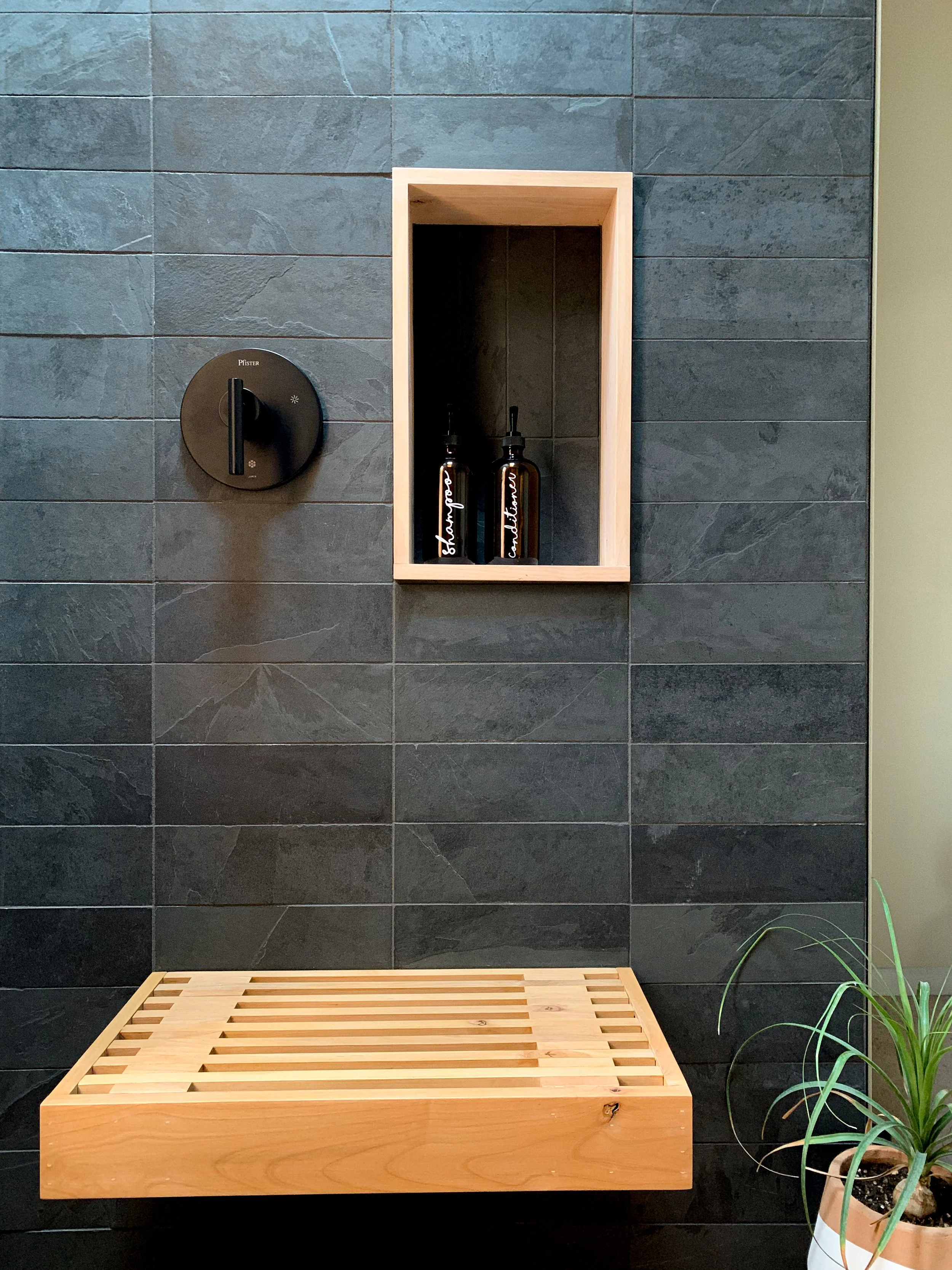 A modern shower area with dark gray tiled wall, a black control knob, a wooden wall niche with two brown bottles of shampoo or conditioner, a wooden slatted bench, and a potted plant in the lower right corner.