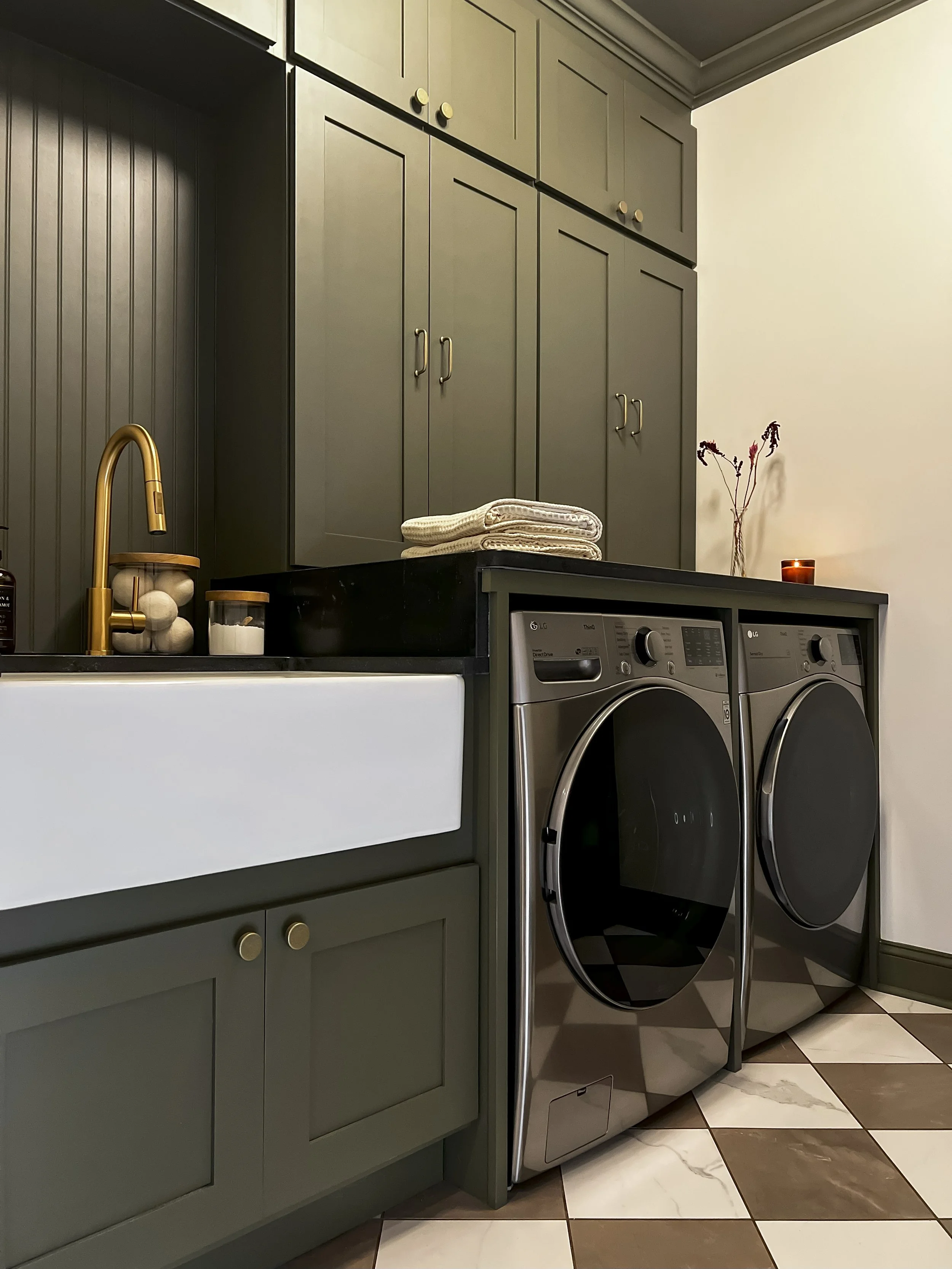 Interior design of an upscale laundry room for a Philadelphia area home. Embodying a modern yet timeless style, this space features dark olive custom cabinets, black granite counter above built-in laundry machines, and checker tile flooring.