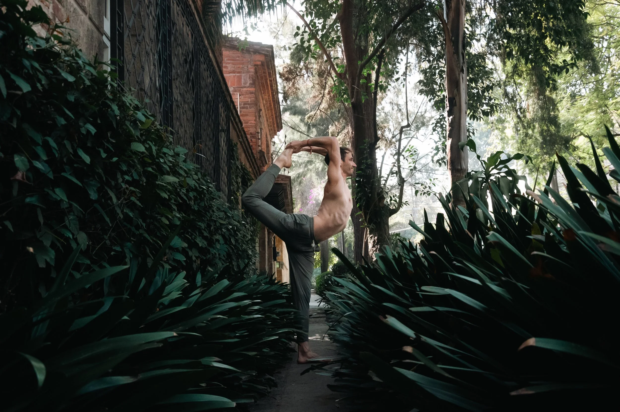 A shirtless man practicing yoga in a street lined with plants and trees, performing a standing backbend with one leg raised behind him and holding his foot with both hands.