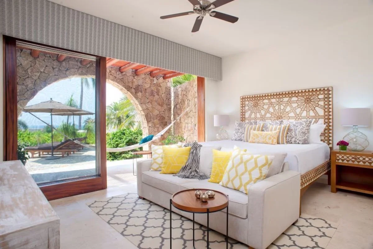 A bedroom with a large bed with patterned headboard, yellow accent pillows, white and grey bedding, and a beige sofa with yellow pillows facing the bed. There is a small round wooden table with metallic decorative balls and a patterned rug. Large sliding glass doors open to an outdoor patio with a hammock, lounge chairs, an umbrella, and tropical greenery.