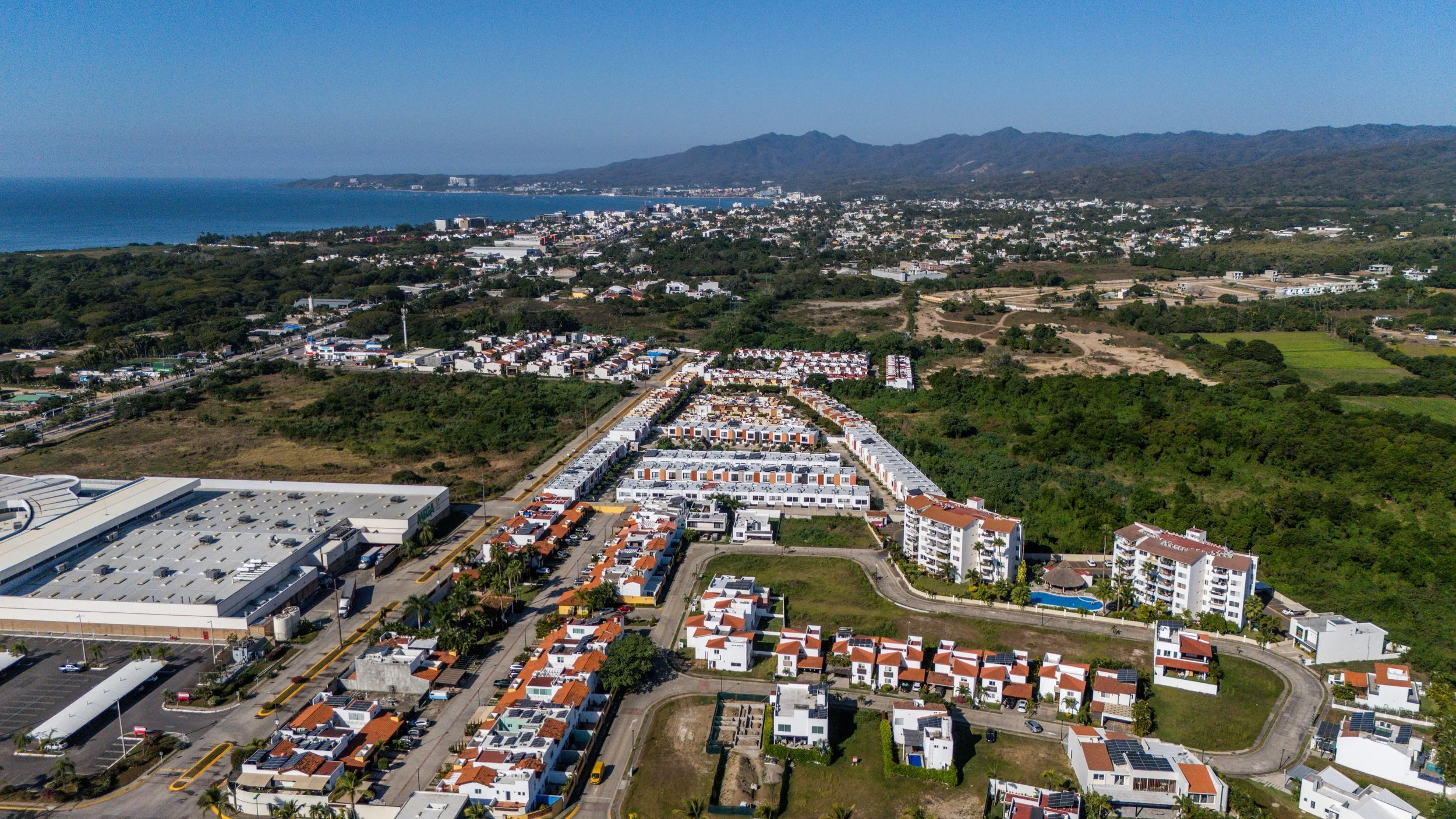 Punta Mita Real Estate Aerial view of a coastal town with residential neighborhoods, commercial buildings, and lush green areas, bordered by a mountain range and a body of water.