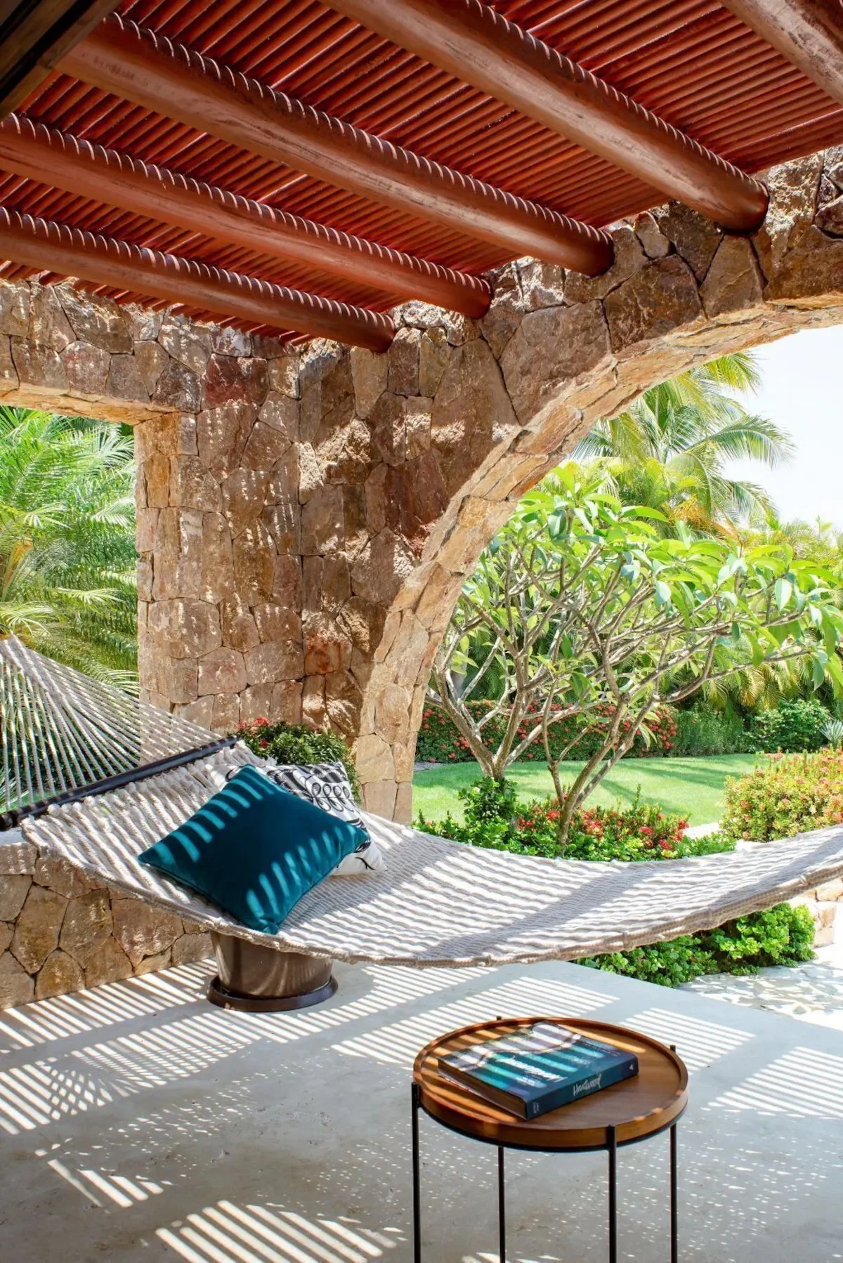 Outdoor patio with a stone wall and arched opening, a hammock with pillows, a small round table with a book, and lush green tropical plants in the background.