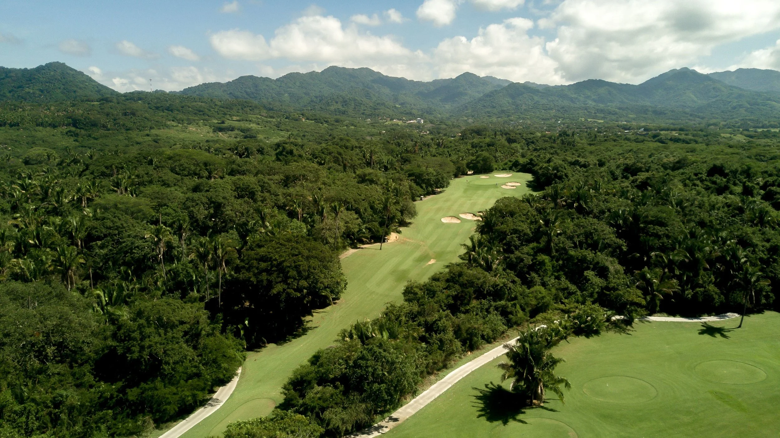 Aerial view of a golf course surrounded by lush green forest and mountains in the background. Punta Mita