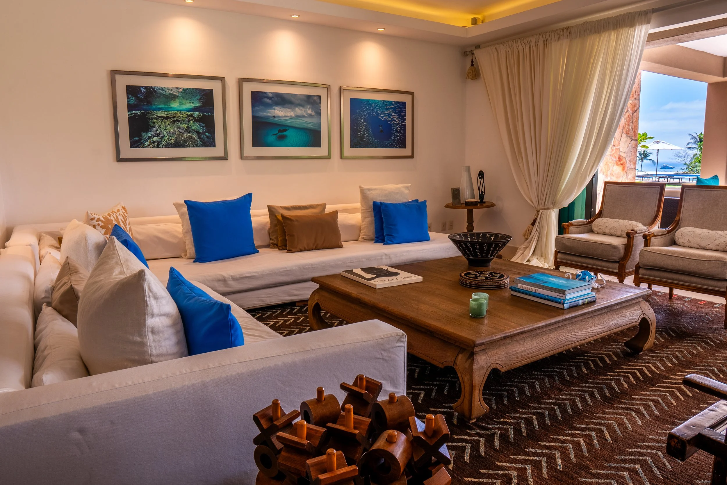 Modern living room with white sofas and blue pillows, wooden coffee table, artworks on the wall, large windows with curtains, tropical outdoor view. Punta Mita Real Estate