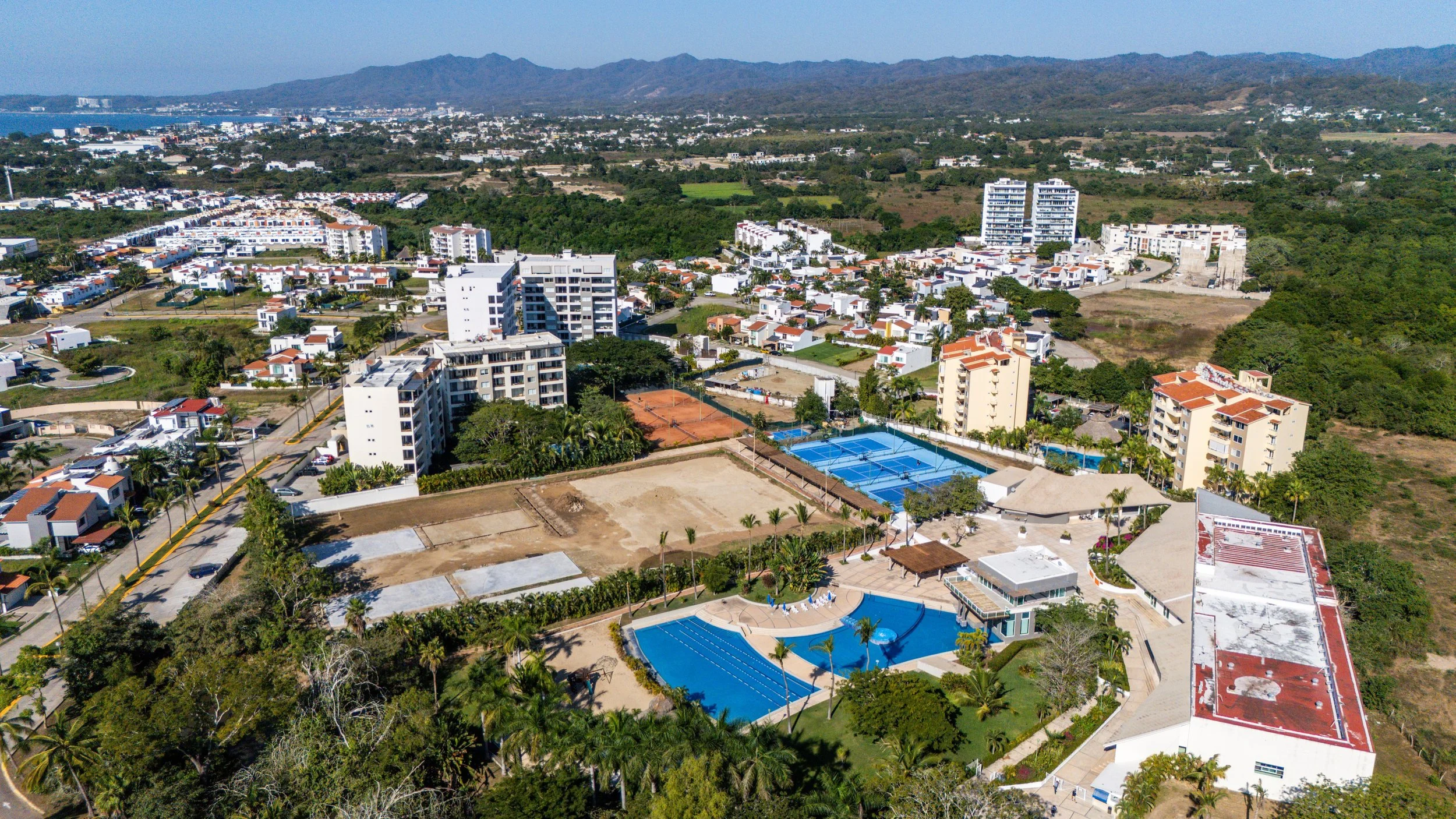 Punta Mita Real Estate Aerial view of a small urban area with residential buildings, tennis courts, and a swimming pool. The background shows mountains and lush greenery.