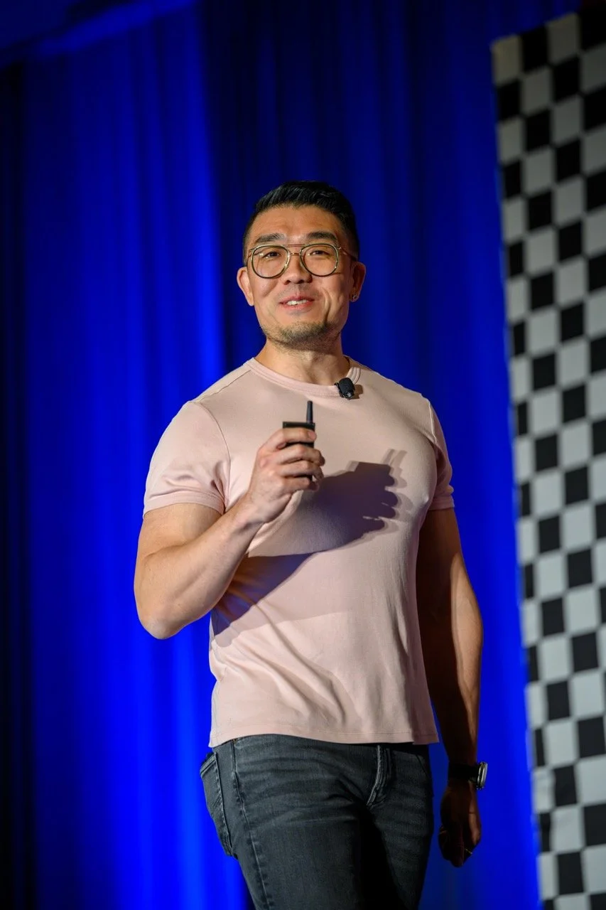 Man with glasses holding a microphone and standing in front of a blue curtain and checkered wall, giving a presentation.