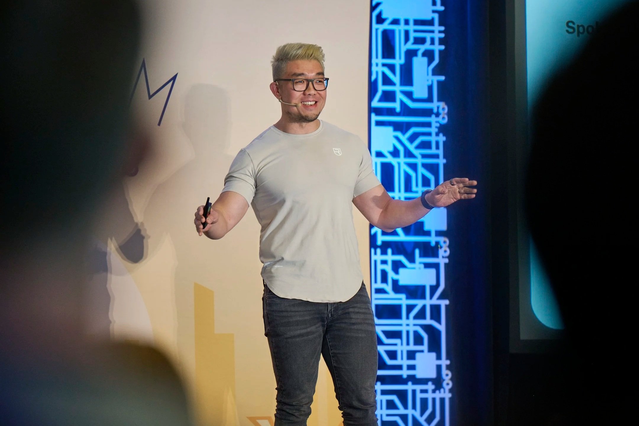 A man with blond hair, glasses, and a gray t-shirt is speaking at a presentation, holding a remote in one hand, with a digital display and a panel of Chinese characters in the background.