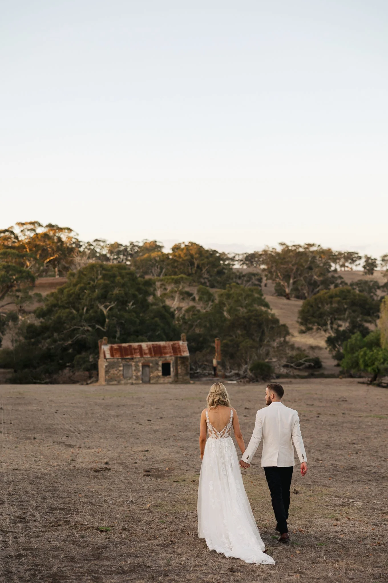 A bride and groom walking hand in hand across a dry open field towards an abandoned rustic building, with trees on a hill in the background and a clear sky.