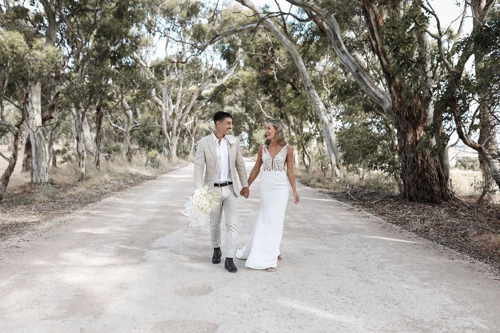 A newlywed couple holding hands and walking down a dirt road surrounded by trees, with the groom holding a bouquet of white flowers.