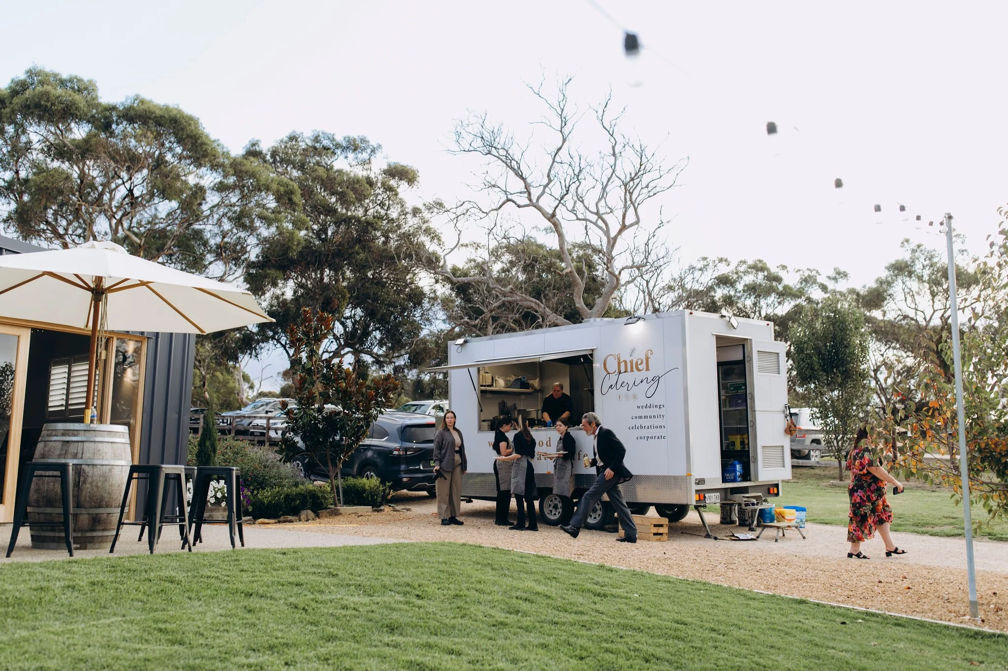 A white catering truck with the sign "Chef Catering" is serving food to a group of people outdoors. There is a large umbrella, some trees, and a grassy area in the background.