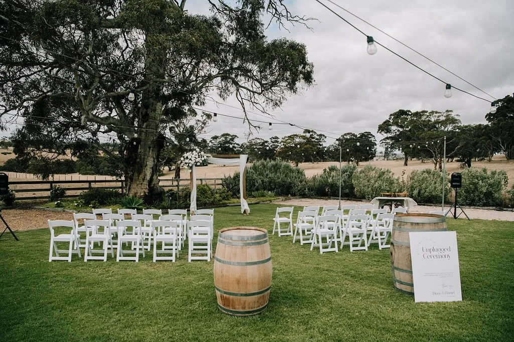 Outdoor wedding ceremony setup with white chairs arranged on a grassy lawn, a wooden arch decorated with flowers, a table on the right, and two wine barrels, with string lights overhead, a tree, and open countryside in the background.