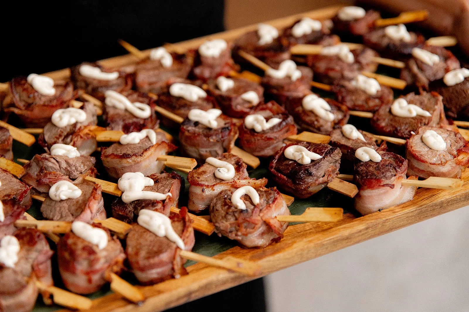 Bite-sized pieces of meat wrapped in bacon, topped with a dollop of white sauce, arranged on a wooden tray.