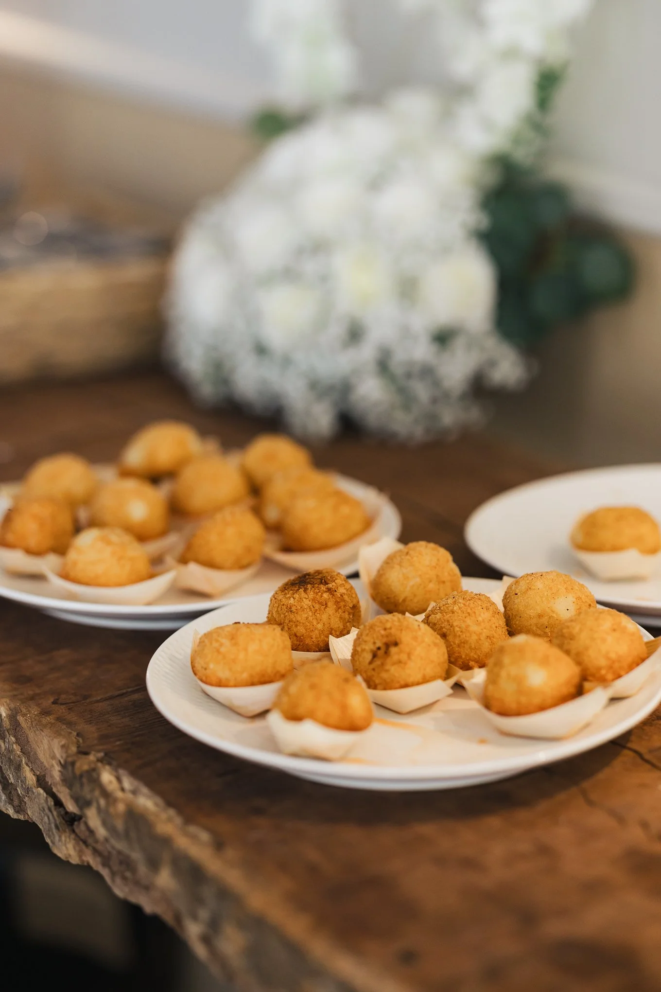 Plate of fried appetizer balls on a rustic wooden table with a blurred bouquet of white flowers in the background.