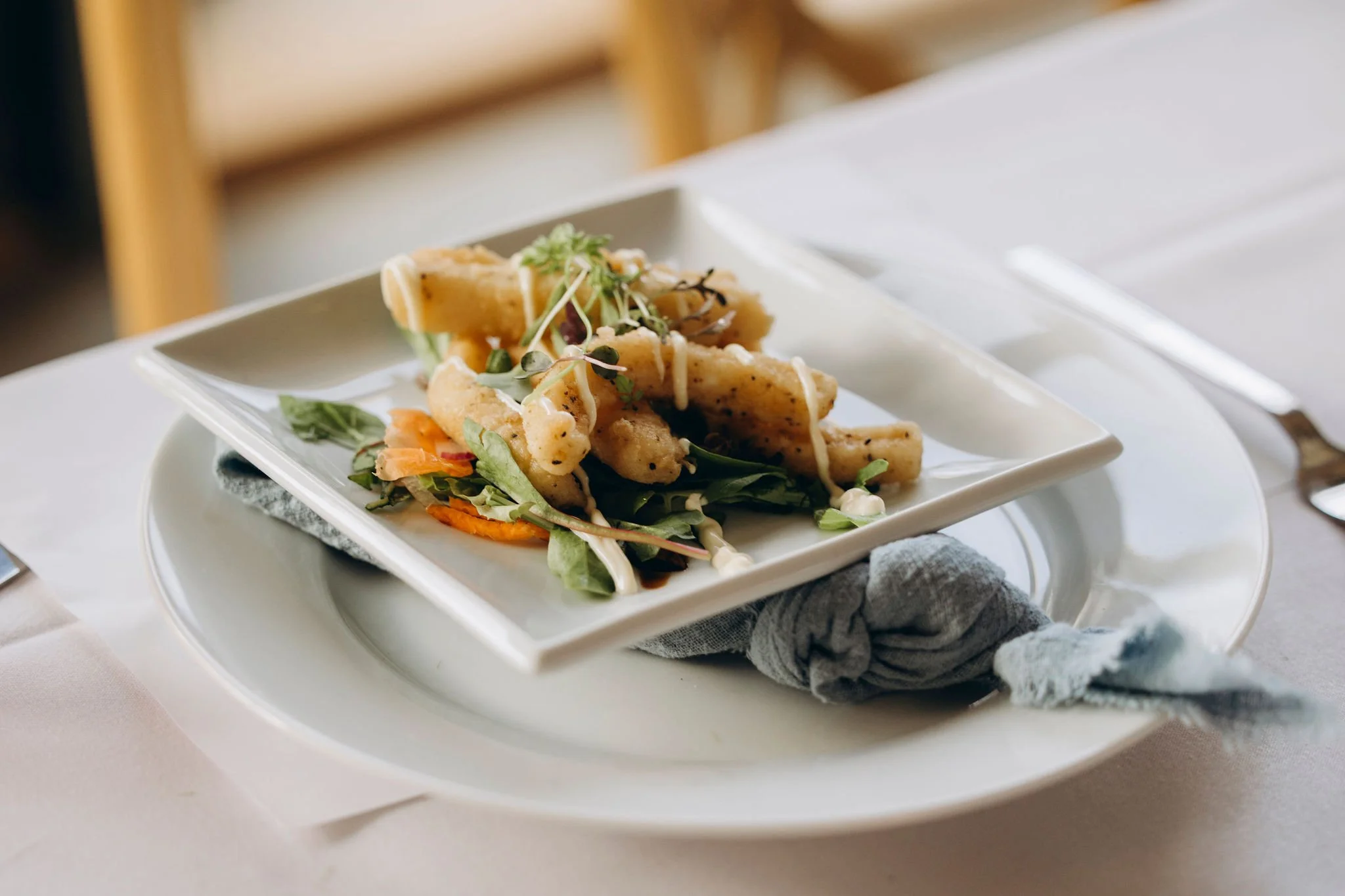 Fried fish on a white square plate, topped with microgreens and drizzled with white sauce, served on a larger white plate with a gray cloth napkin underneath.