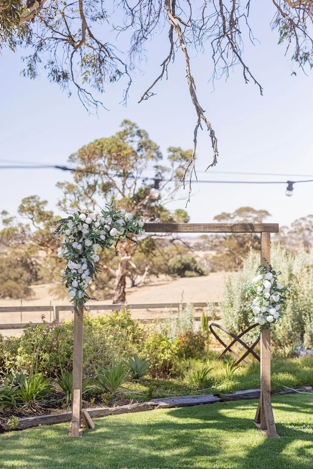 Outdoor wedding arch decorated with white flowers and greenery, set in a garden area with trees and a grassy lawn.