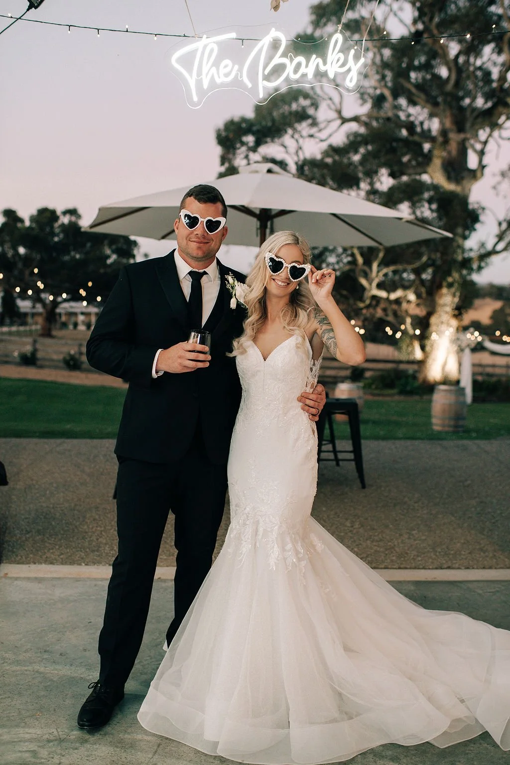 A bride and groom at their wedding reception, both wearing heart-shaped sunglasses and smiling, standing under an umbrella with string lights and a neon sign that says 'The Banks' in the background.