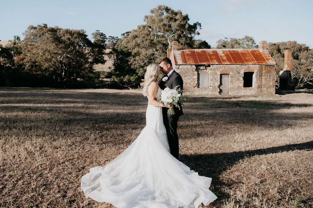 A bride and groom on their wedding day, standing close and holding a bouquet of flowers outdoors in front of an old house with a rusted metal roof.