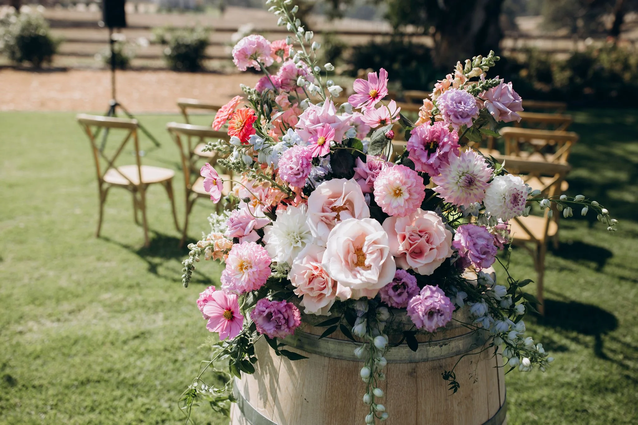 A large bouquet of pink, purple, and white flowers in a wooden barrel on a grassy outdoor setting with empty chairs in the background.