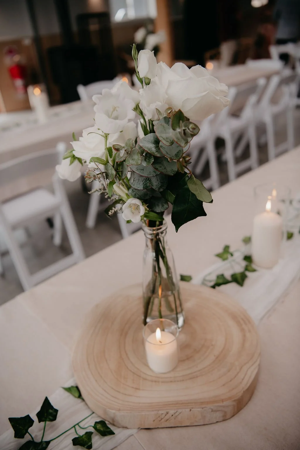A floral centerpiece with white flowers and green leaves in a clear glass vase, placed on a wooden slab, with candles and greenery on a beige tablecloth at a decorated event venue.
