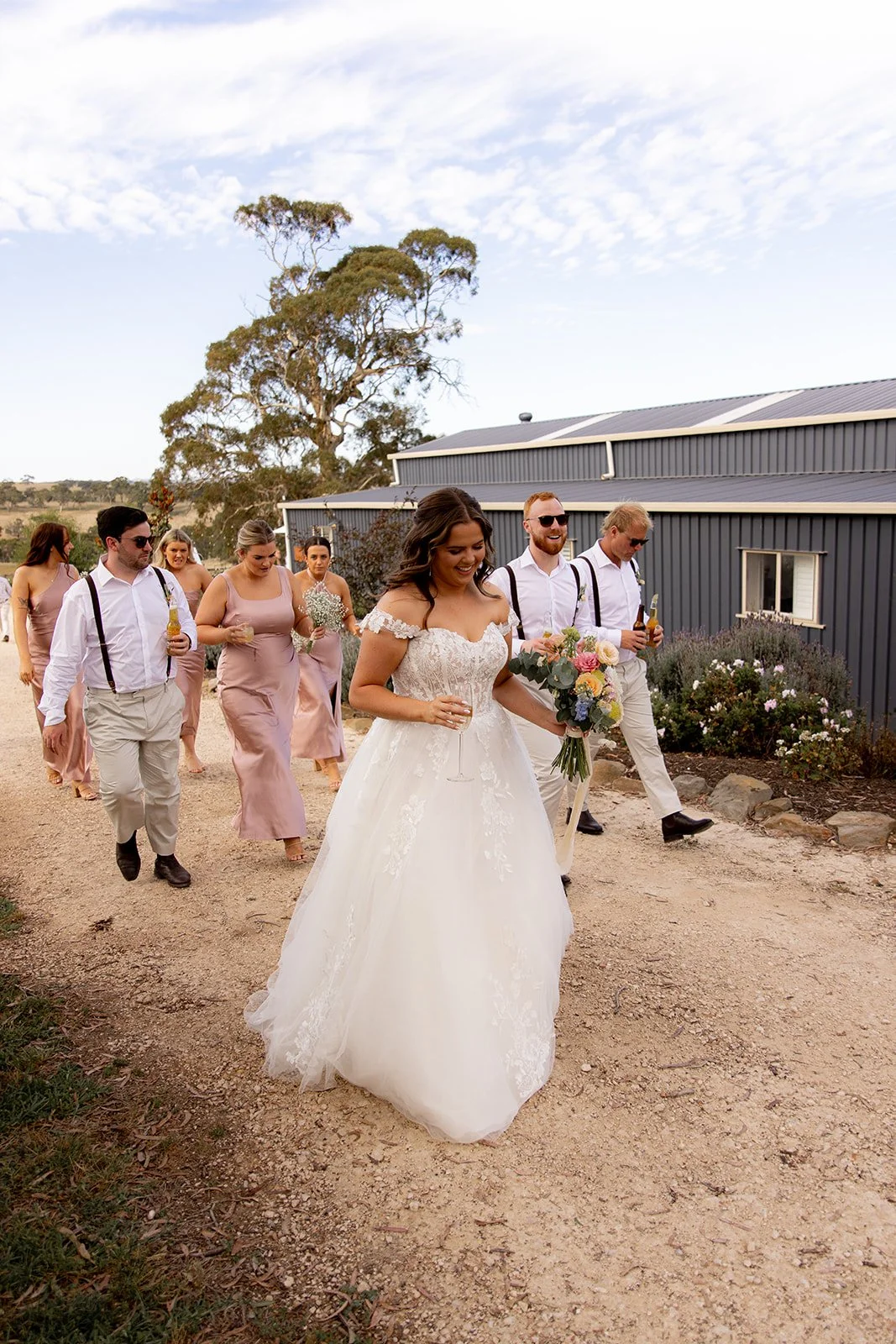 A bride in a white wedding dress holding a bouquet and a glass of champagne, walking outdoors with her wedding party including bridesmaids and groomsmen.