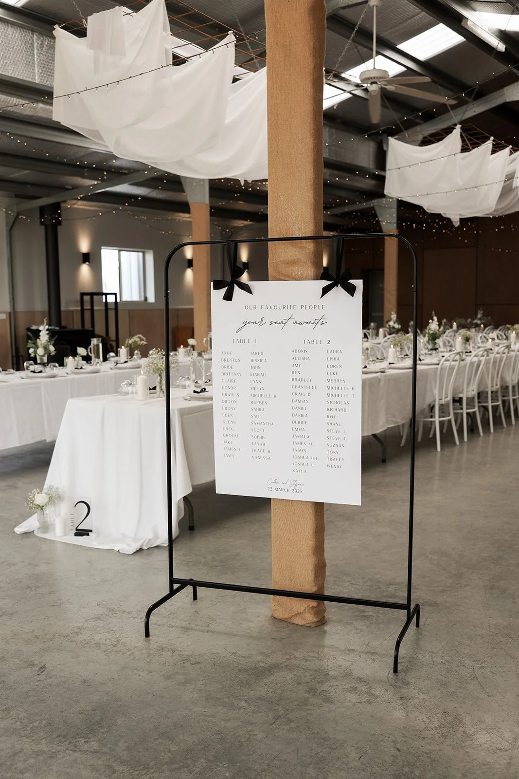 Wedding reception with long tables decorated with white tablecloths, flowers, and candles. A seating chart sign with black bows hanging from a metal stand shows the list of guests and table assignments, with a modern industrial-style venue featuring