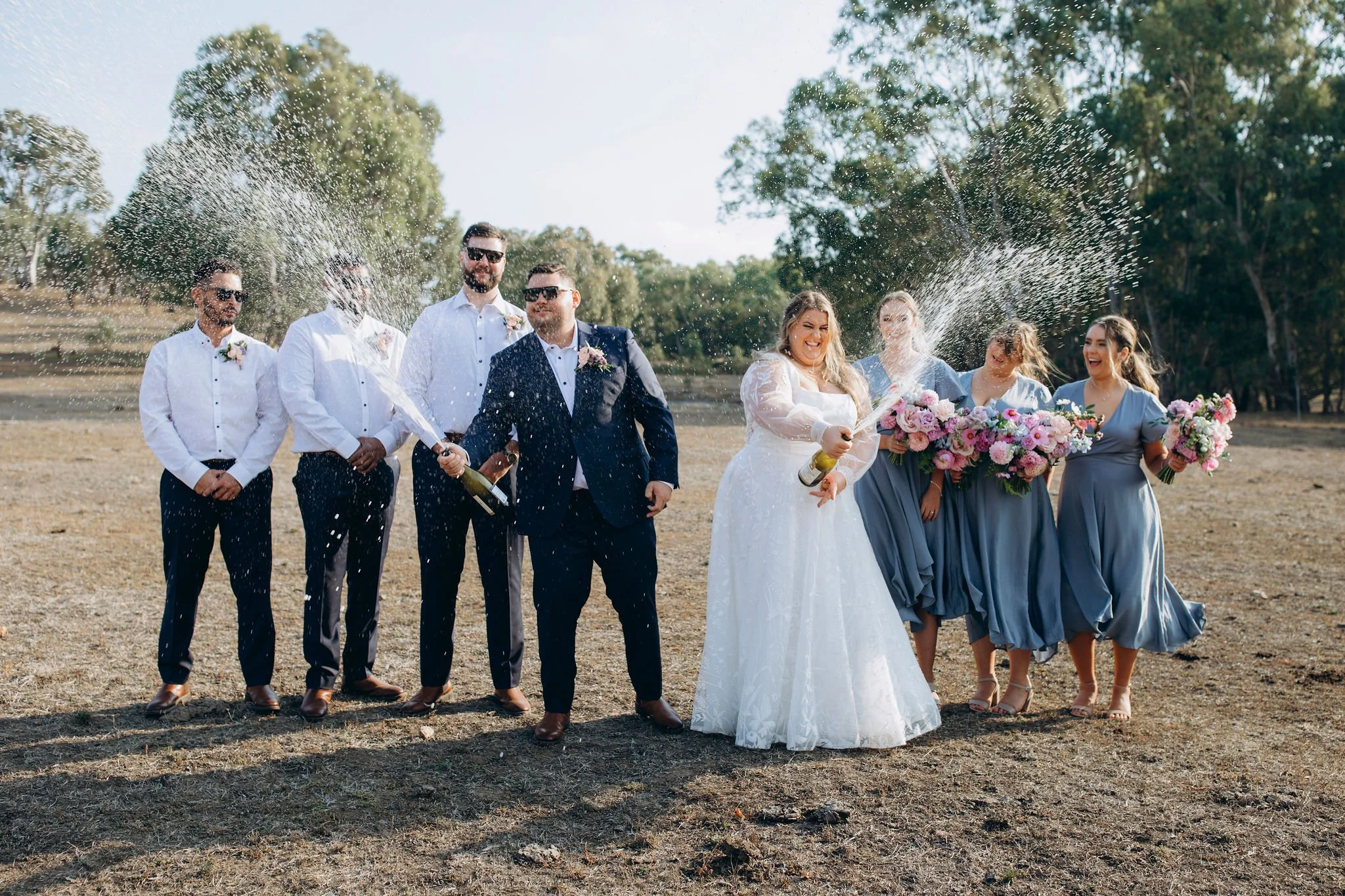 A wedding celebration outdoors with seven people. The groom and groomsmen are dressed in dark suits and white shirts. The bride and bridesmaids wear light blue dresses, holding bouquets of pink flowers. Some of the group are laughing while spraying c