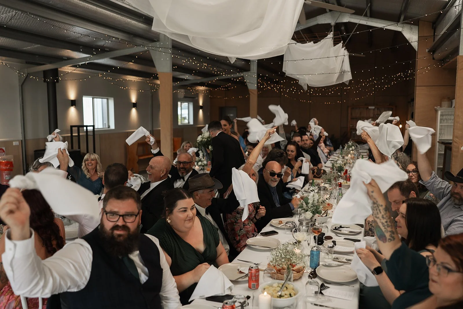 Guests at a wedding reception waving napkins in celebration, seated around a long decorated table indoors with string lights overhead.