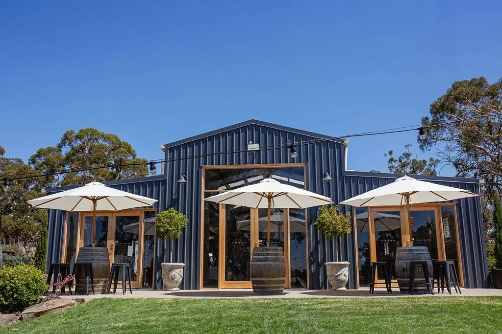 Exterior of a modern building with a metal facade, large glass windows, and outdoor seating with umbrellas in a landscaped setting.