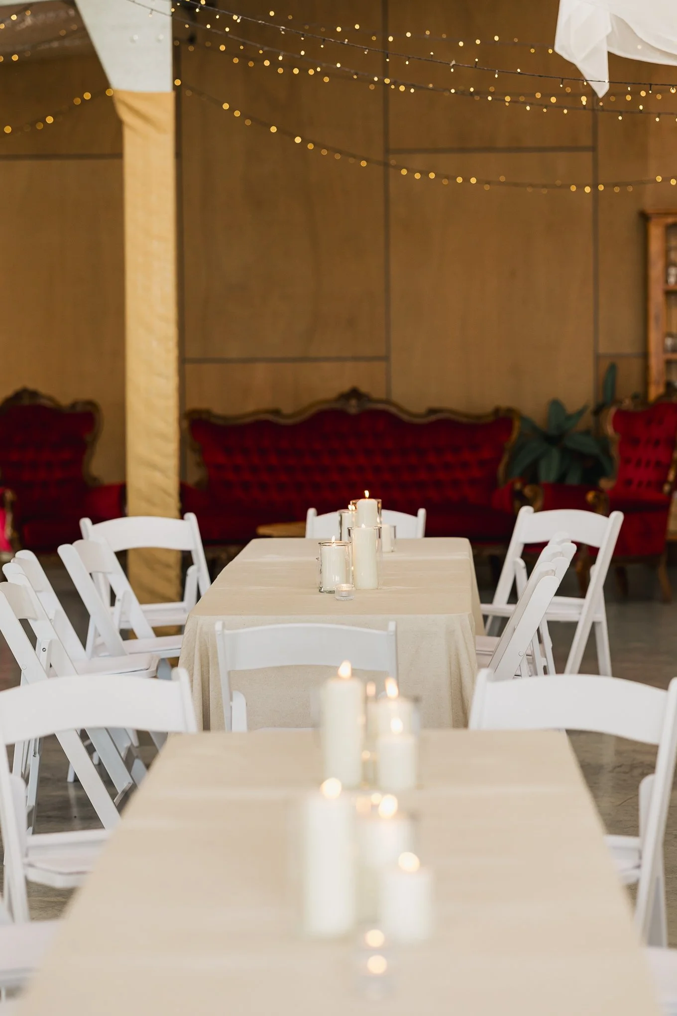 Empty banquet hall with white tables, white chairs, candles, and string lights, with red velvet sofas in the background.