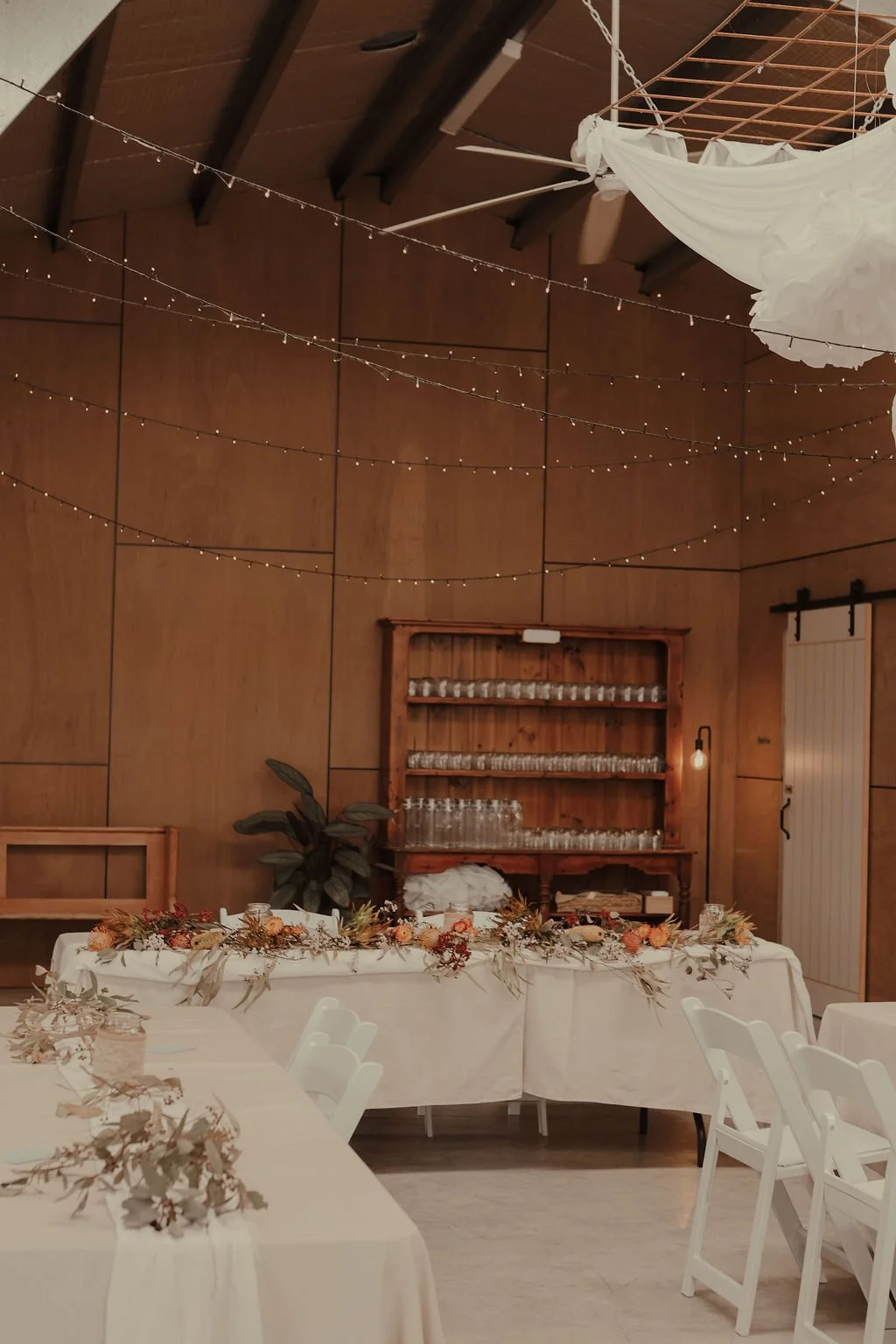 Event space decorated with string lights, a floral table runner, and wooden accents including a shelf with glassware and a barn door.
