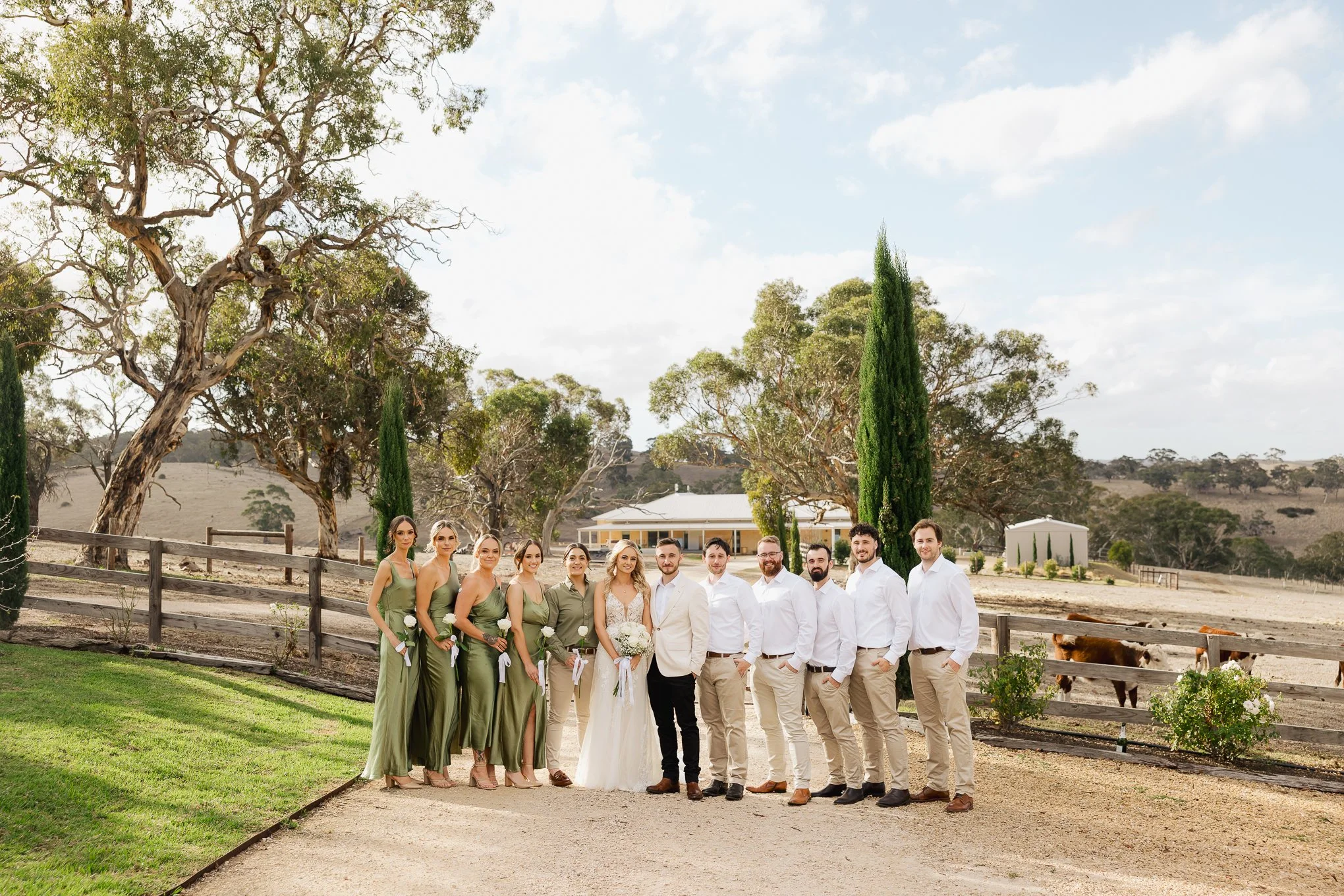 A wedding party of nine people standing outdoors in a rural setting with trees, a fence, and a barn in the background. The bride and groom are in the center, dressed in a white gown and light-colored suit, respectively. Four bridesmaids in green dres