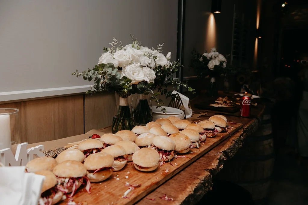 A wooden table with a line of small sandwiches, a vase of white roses, a candle, a can of Coca-Cola, and a sign that says 'Mrs.', at a celebratory event.