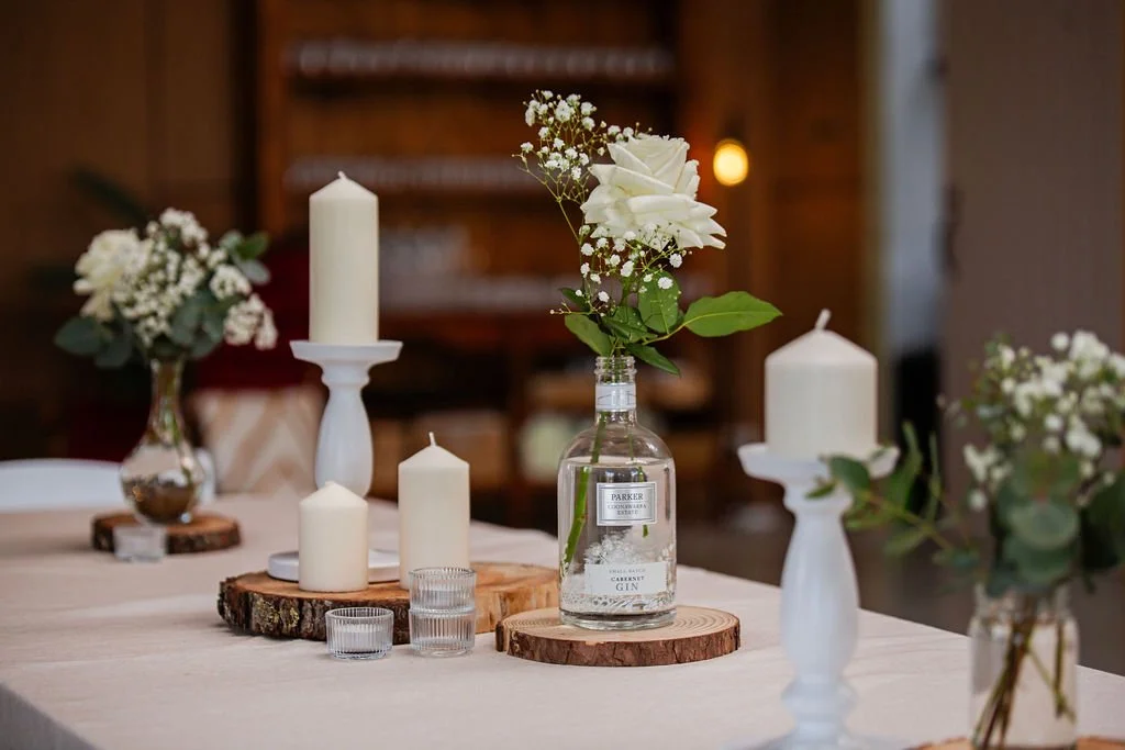 Decorative table setting with white candles, white roses, and greenery in glass vases on wooden slabs, in a cozy indoor space.