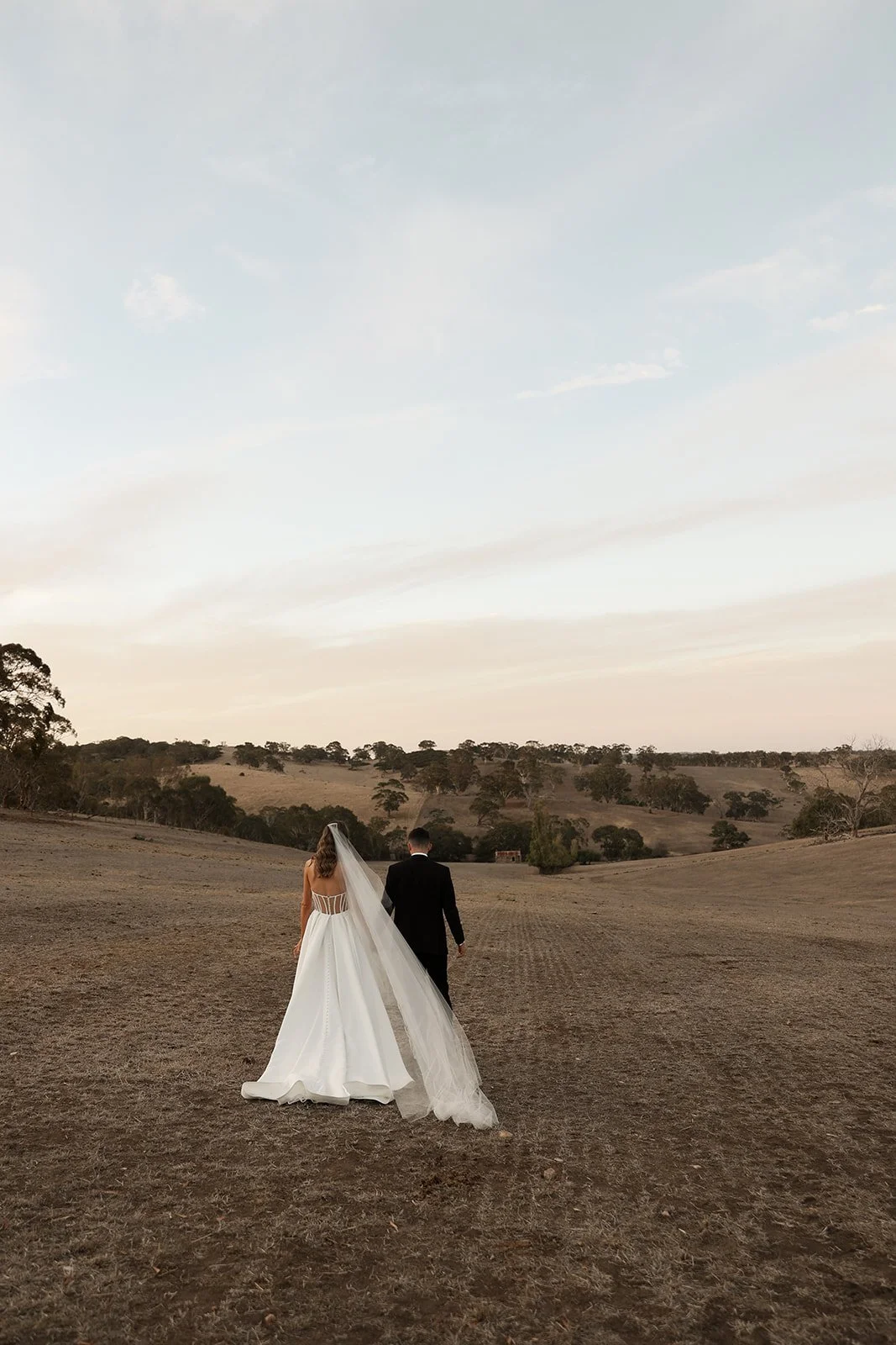 A bride in a white wedding gown and a groom in a black suit walking away in a vast open field at sunset.
