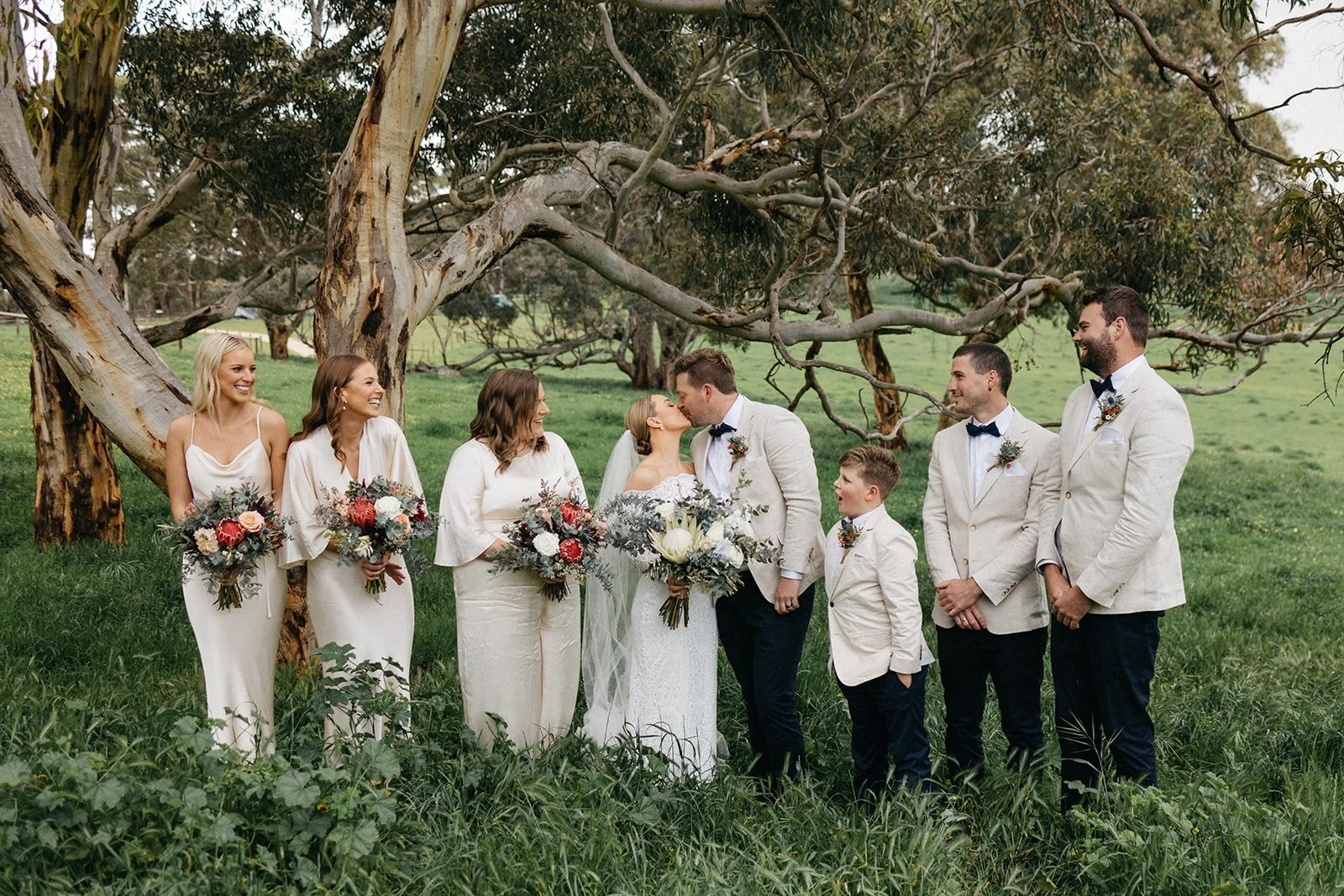 A wedding party outdoors with the bride and groom kissing, surrounded by bridesmaids and groomsmen, under large trees on green grass.