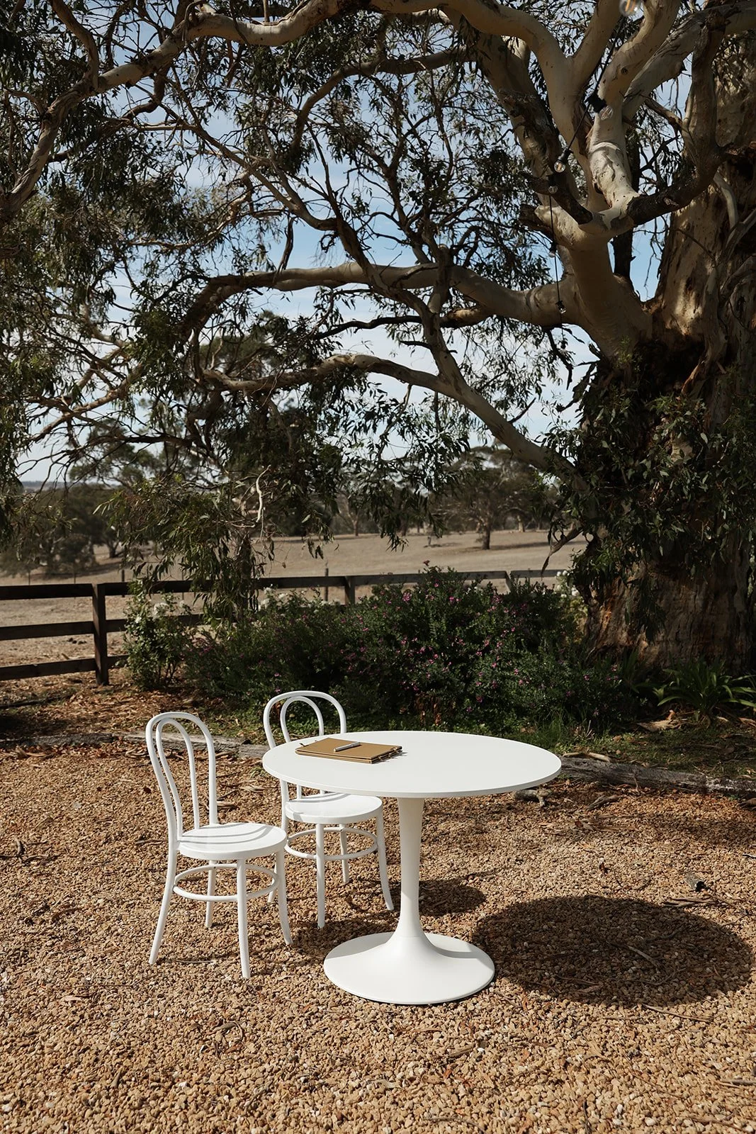 A white round table with two white chairs beside it, set outdoors on a gravel surface with a large tree and some bushes in the background.