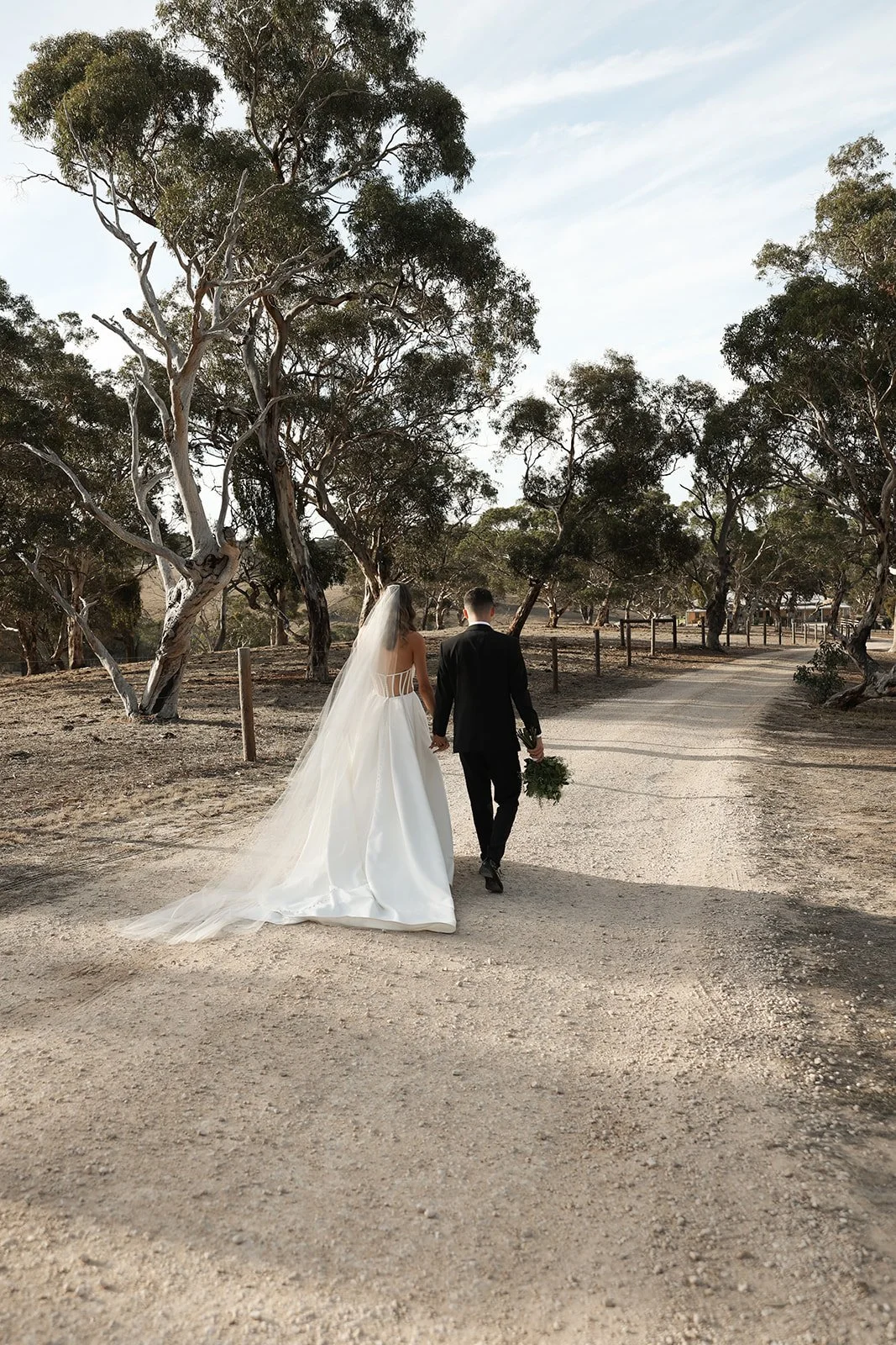 A bride and groom holding hands and walking along a dirt path lined with trees, with the bride in a white wedding dress and veil, and the groom in a black suit, holding a bouquet of greenery.