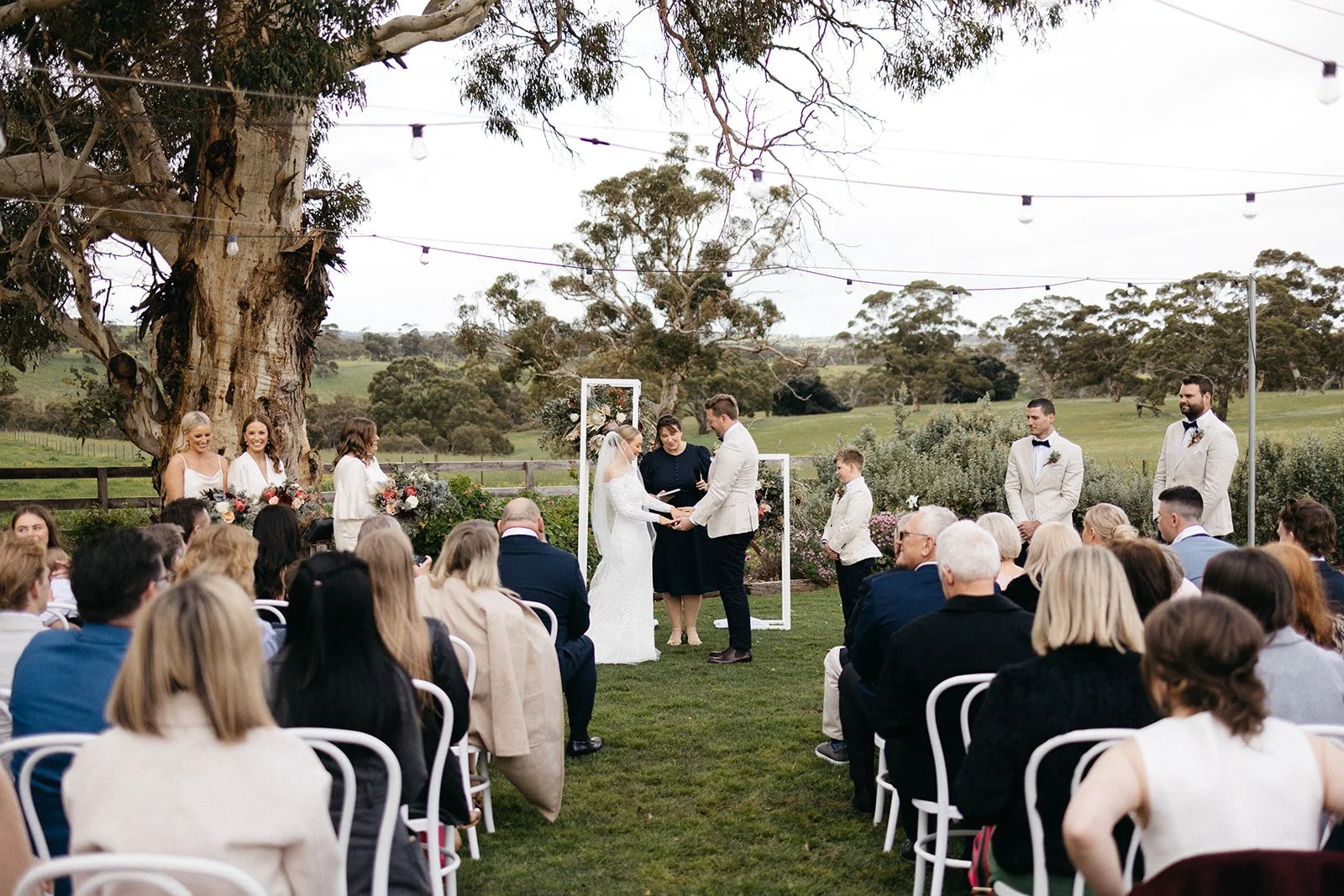 An outdoor wedding ceremony with a couple exchanging vows under an arch, surrounded by guests seated on white chairs, with a large tree and rolling green hills in the background.
