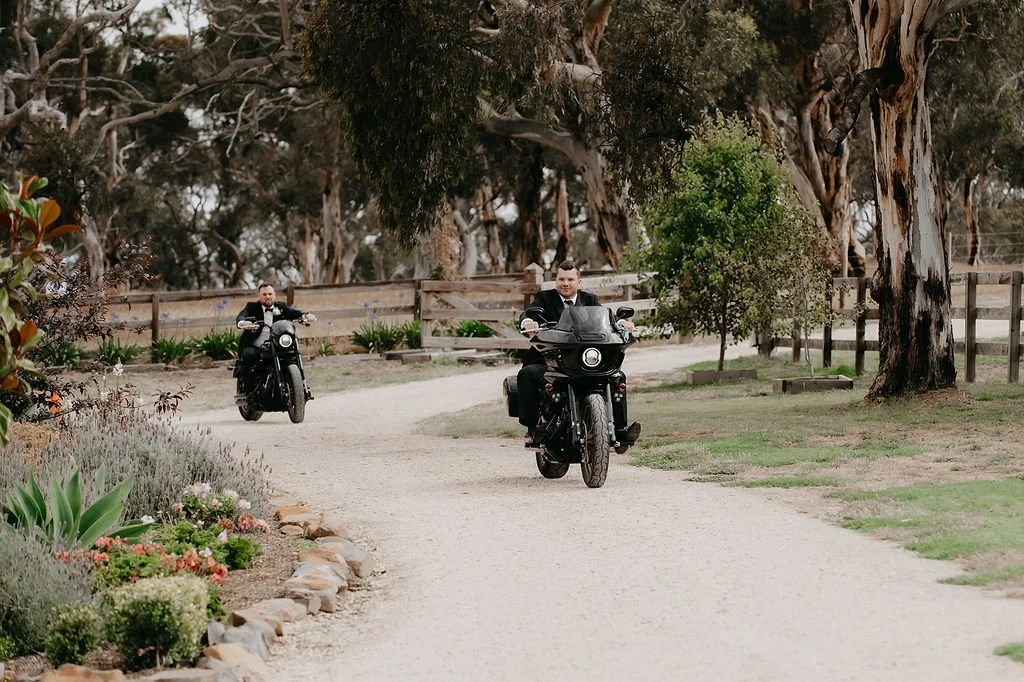 Two men riding black motorcycles along a dirt path in a park with trees and plants.