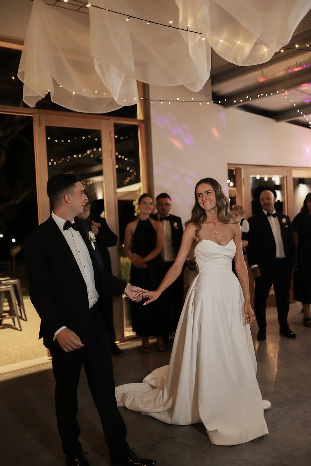A bride and groom holding hands during their wedding reception, surrounded by guests in formal attire, with elegant decorations and lighting overhead.