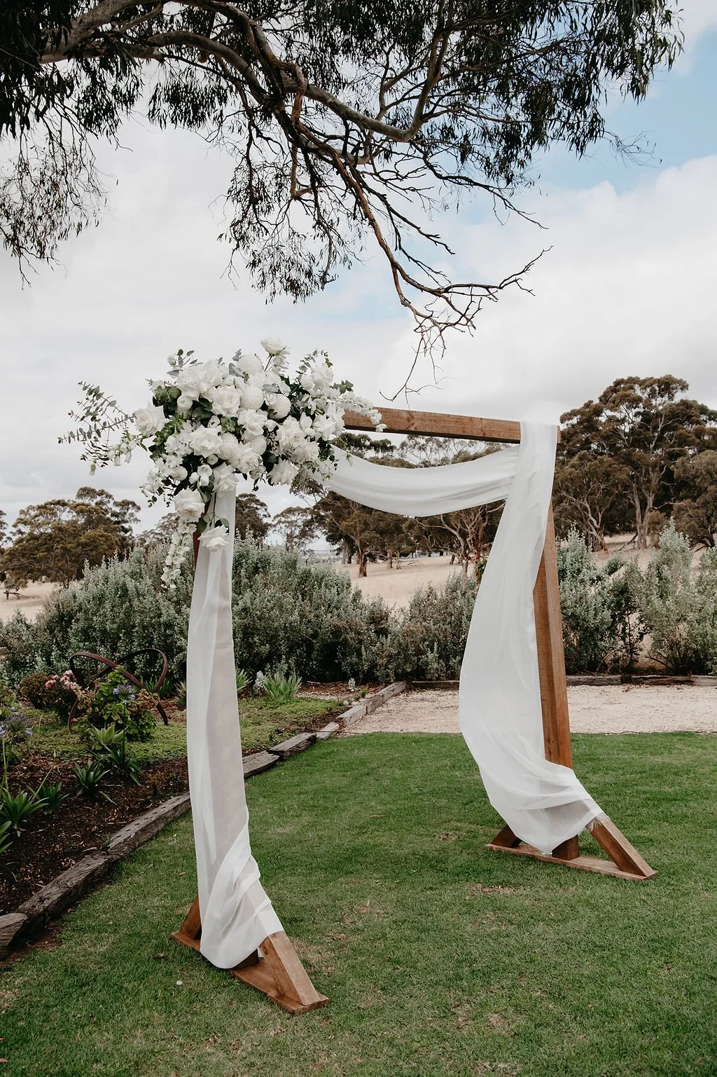 Outdoor wedding arch decorated with white flowers and flowing white fabric, set on a grassy area with garden and trees in the background.