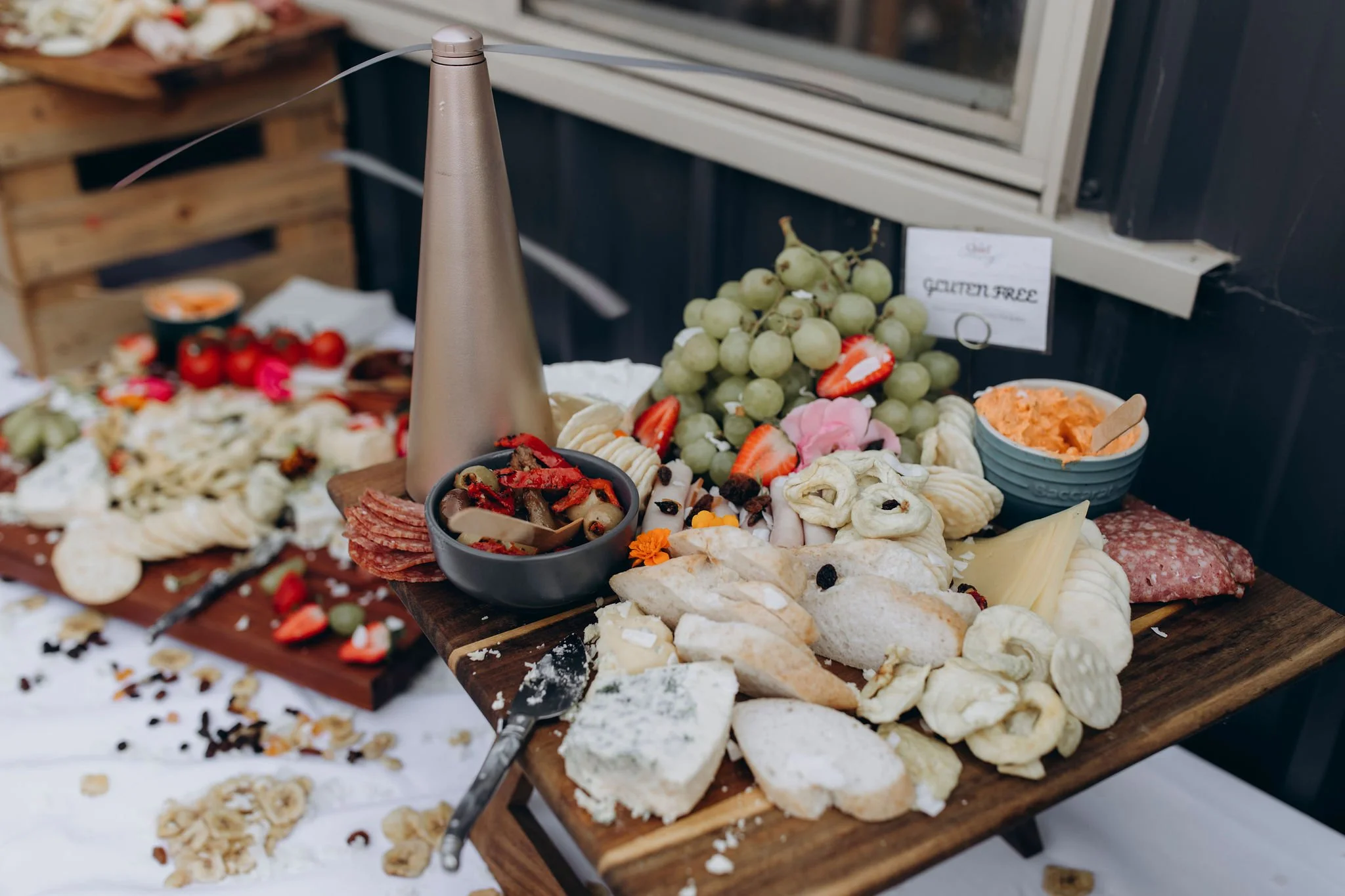 Cheese, grapes, strawberries, cured meats, bread, and dips on a wooden cheese board, with a bottle of wine or champagne on a table.