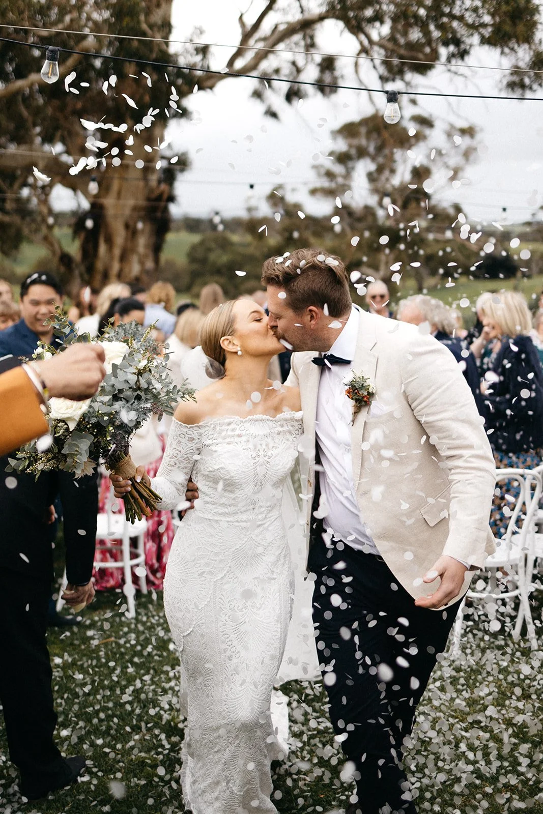 A newlywed couple shares a kiss at their outdoor wedding ceremony, surrounded by guests, confetti, and string lights under a large tree.