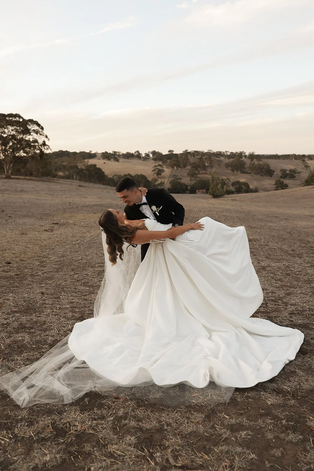 A newlywed couple in wedding attire enjoying a moment together in an open, rural landscape at sunset. The groom is dressed in a black tuxedo and is holding the bride, who is in a white wedding gown, as they smile at each other.