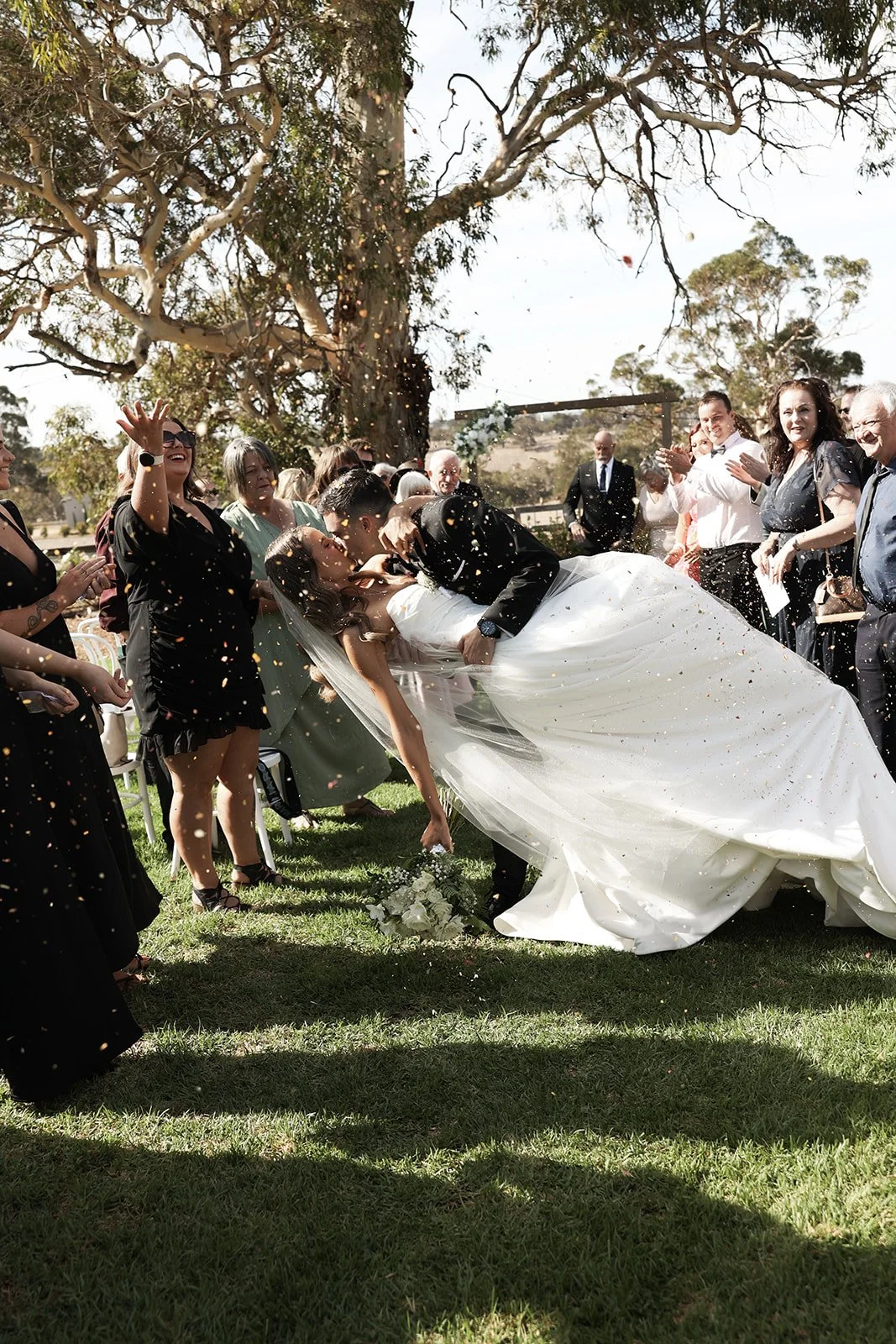A bride and groom sharing a kiss while surrounded by smiling guests at an outdoor wedding ceremony, with confetti and trees in the background.